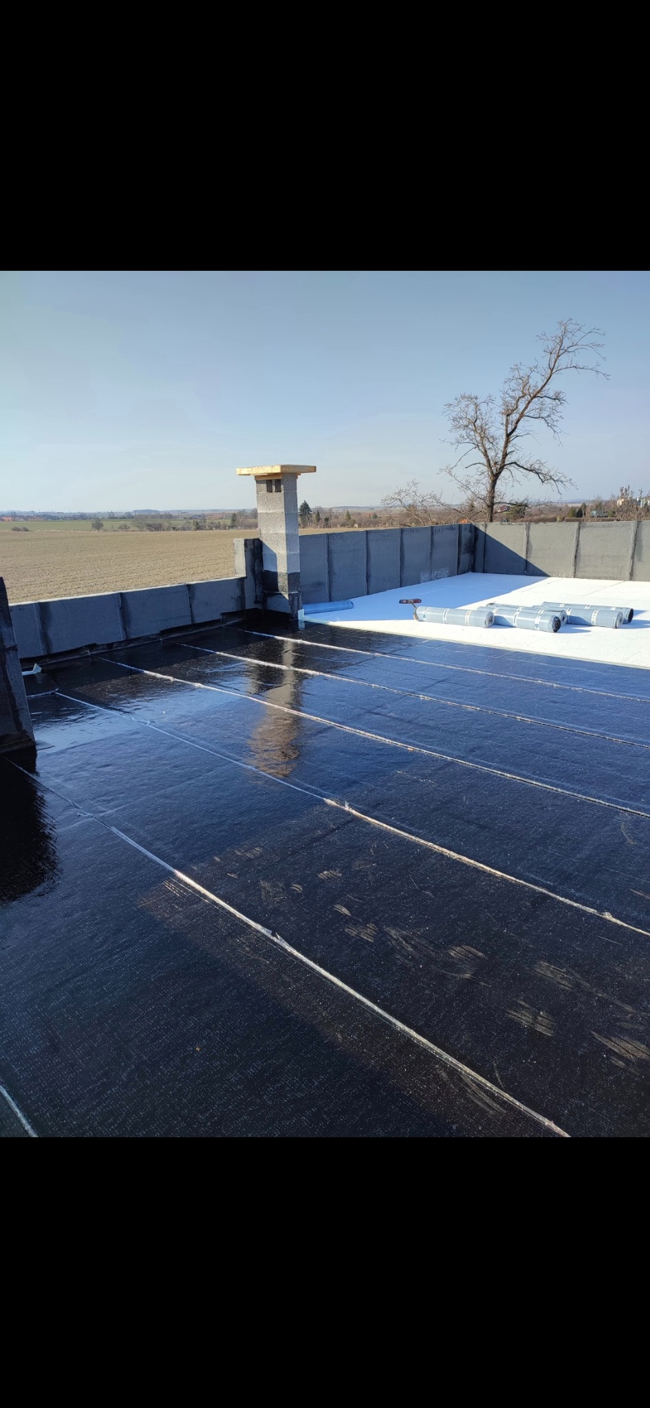 Płaski dach pokryty czarną papą termozgrzewalną, z widocznym kominem i rolkami papy w tle na tle błękitnego nieba i krajobrazu.