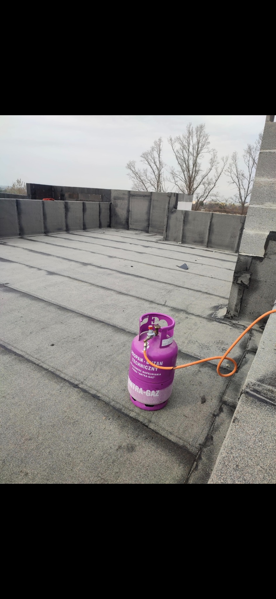 Prace dekarskie na dachu płaskim: widoczna butla gazowa z podłączonym przewodem na tle pokrycia papą. W oddali elementy konstrukcyjne dachu i drzewa bez liści.
