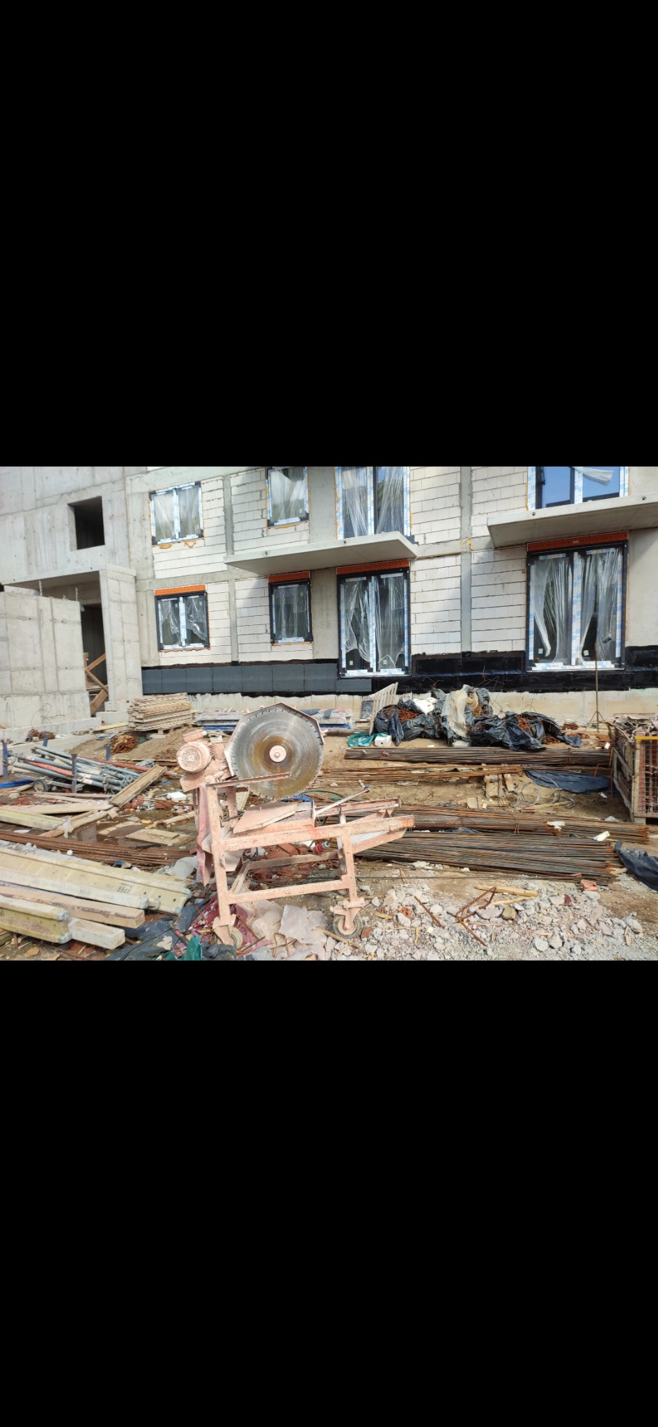 Budynek w trakcie budowy z widoczną piłą tarczową, elementami konstrukcyjnymi i zabezpieczonymi oknami. Ujęcie z bliska na placu budowy.