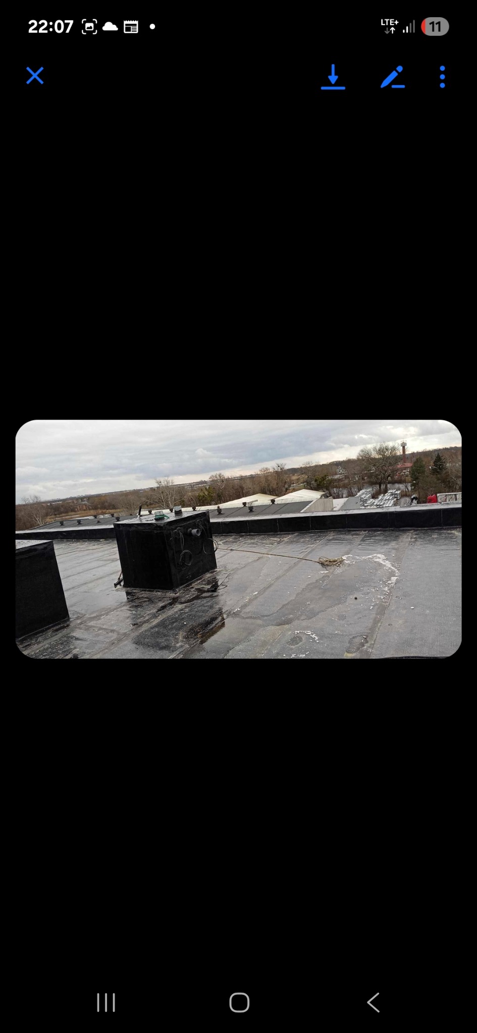 Płaski dach pokryty papą z widocznymi elementami wentylacyjnymi i zbiornikami, po deszczu. W tle zabudowania i drzewa pod pochmurnym niebem.