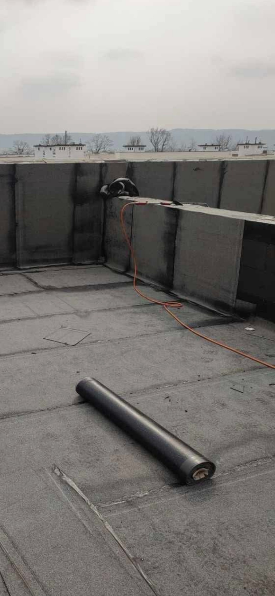 Dekarz pracuje na płaskim dachu, widoczna rolka papy, zgrzewarka i pomarańczowy kabel. W tle budynki i pochmurne niebo.