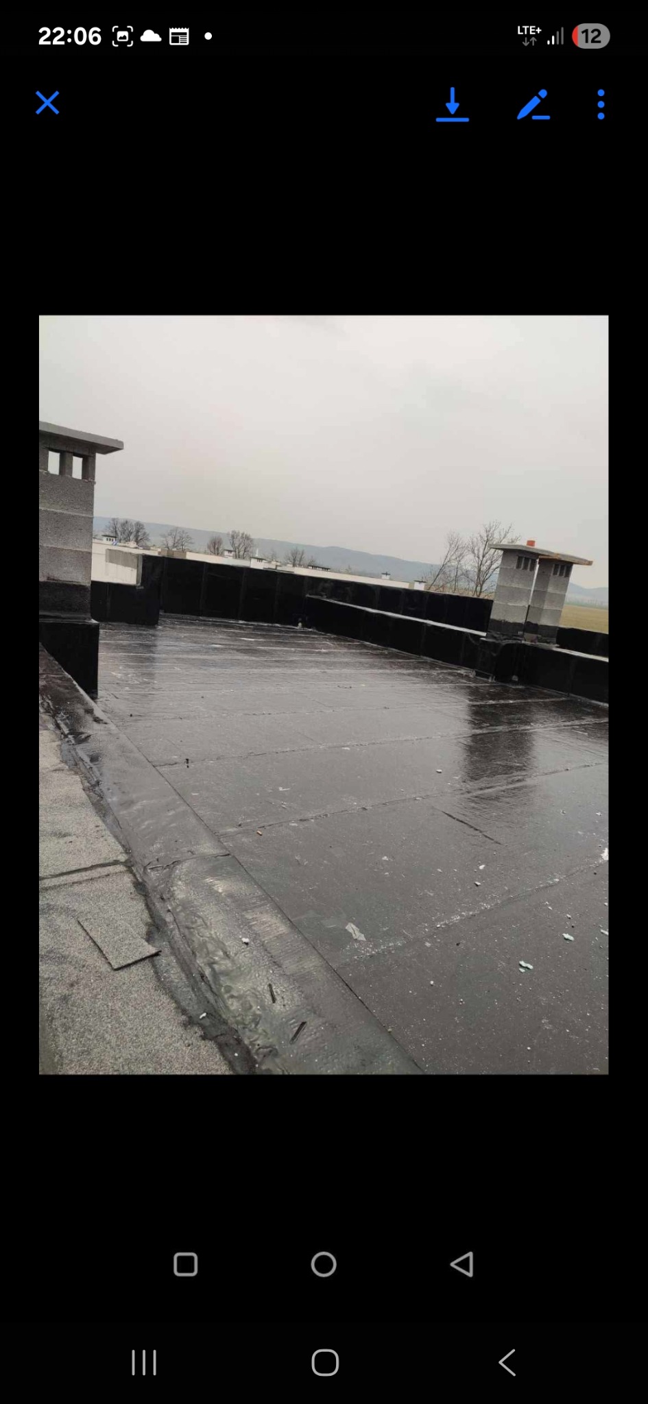 Wykończony, płaski dach pokryty czarną papą bitumiczną z dwoma kominami z szarego pustaka. Widok na okoliczne pola i lasy w pochmurny dzień.