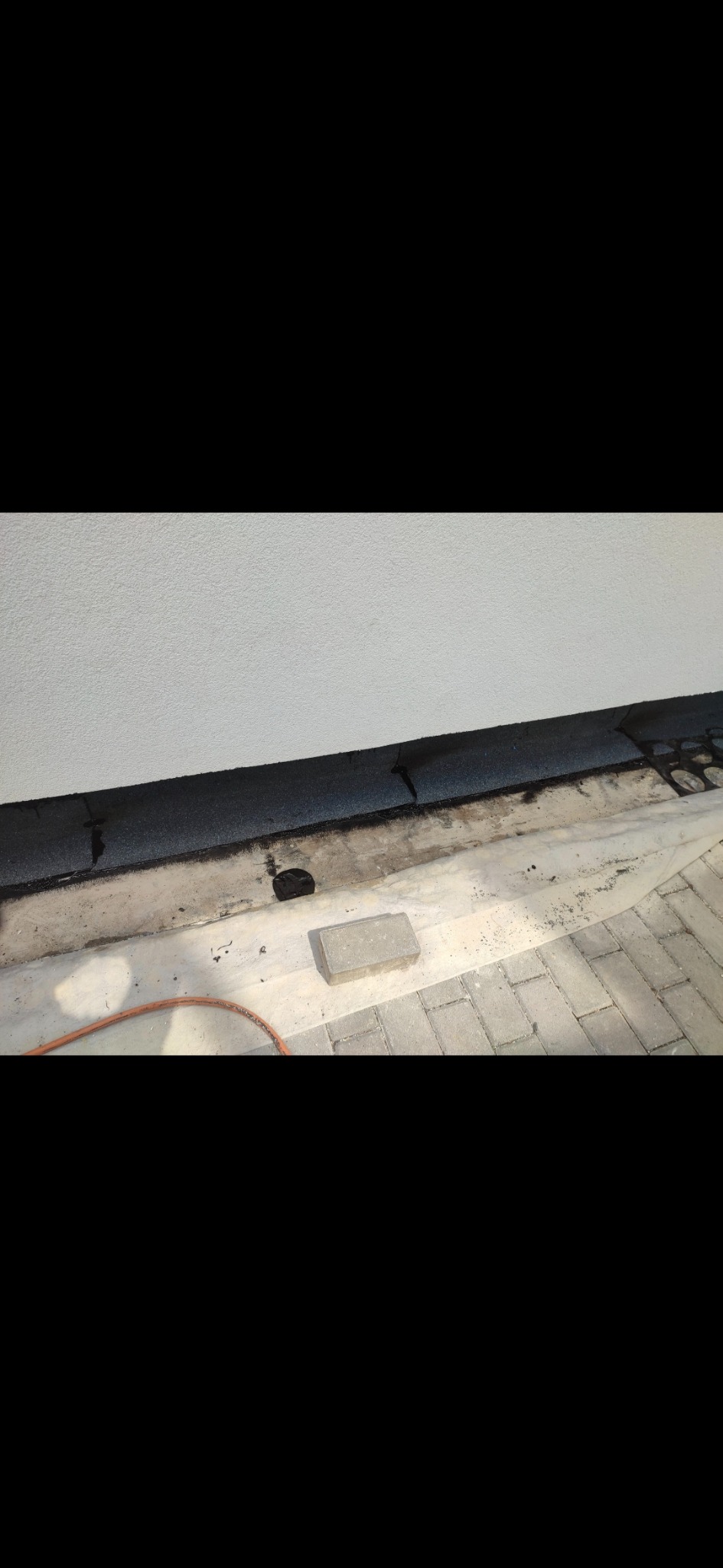 Detal dachu przy ścianie budynku po uszczelnieniu. Widoczne elementy papy, cegła i kostka brukowa. Praca w trakcie realizacji, z bliska.