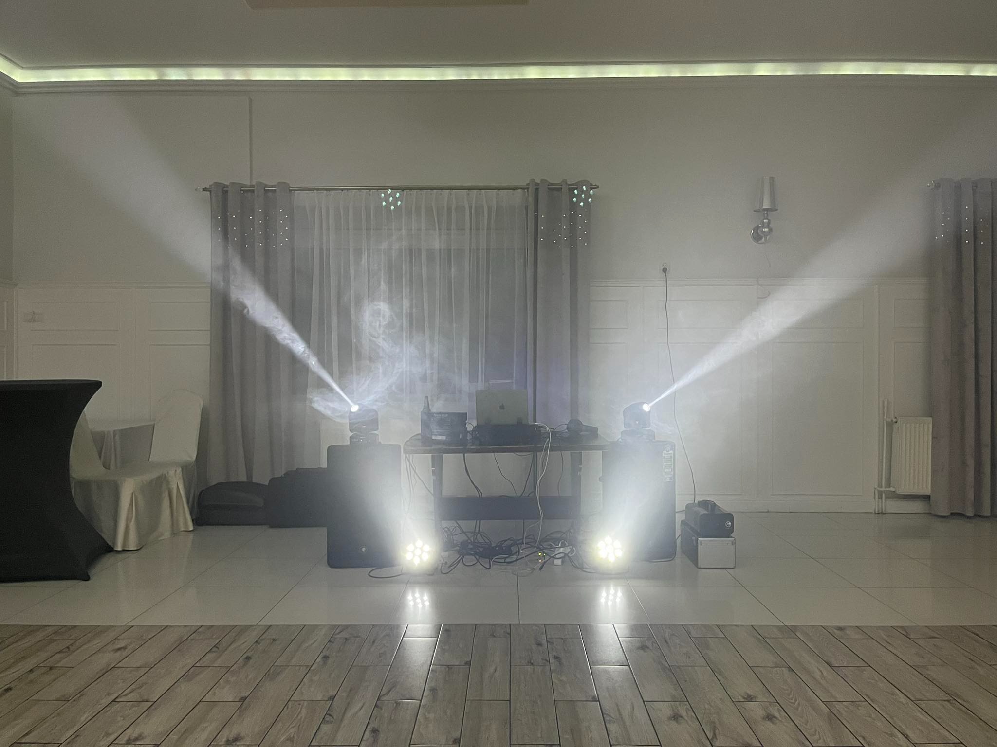 Scena z nagłośnieniem i oświetleniem eventowym. Reflektory tworzą efektowne snopy światła. Widoczny sprzęt DJ-ski i głośniki. Parkiet w odcieniach brązu i beżu.