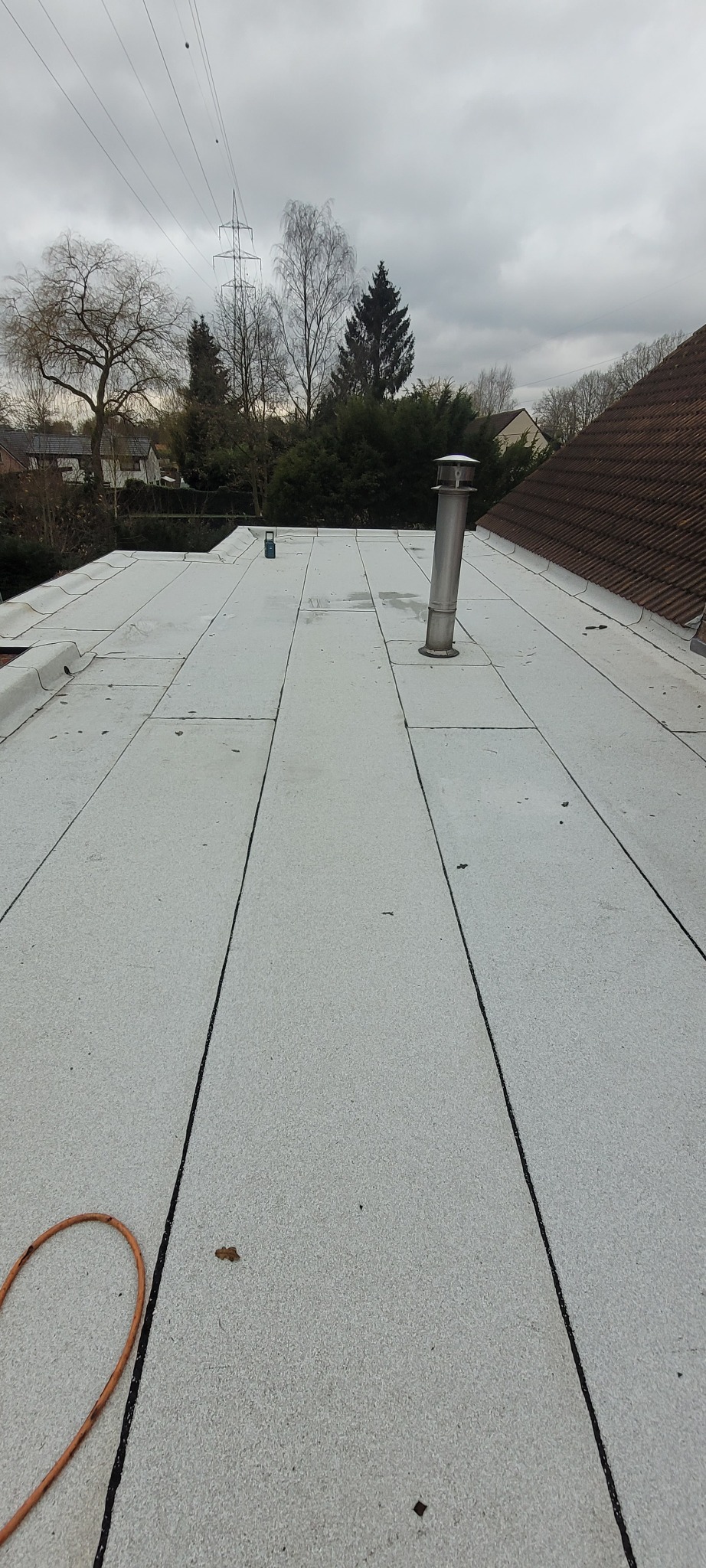 Płaski dach pokryty jasnymi panelami z widocznym kominem i narzędziem, w tle drzewa i pochmurne niebo.