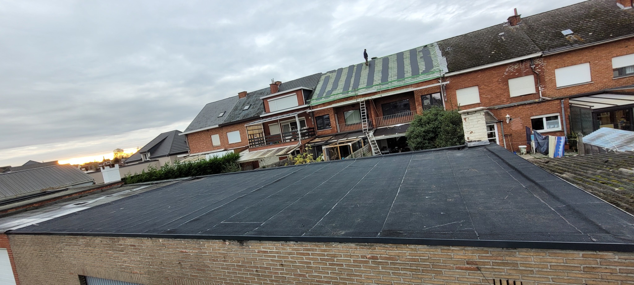 Widok z góry na kilka domów z różnymi rodzajami pokrycia dachowego, w tym jeden z czarną papą termozgrzewalną i jeden w trakcie wymiany z widocznymi łatami i pracownikiem na dachu.