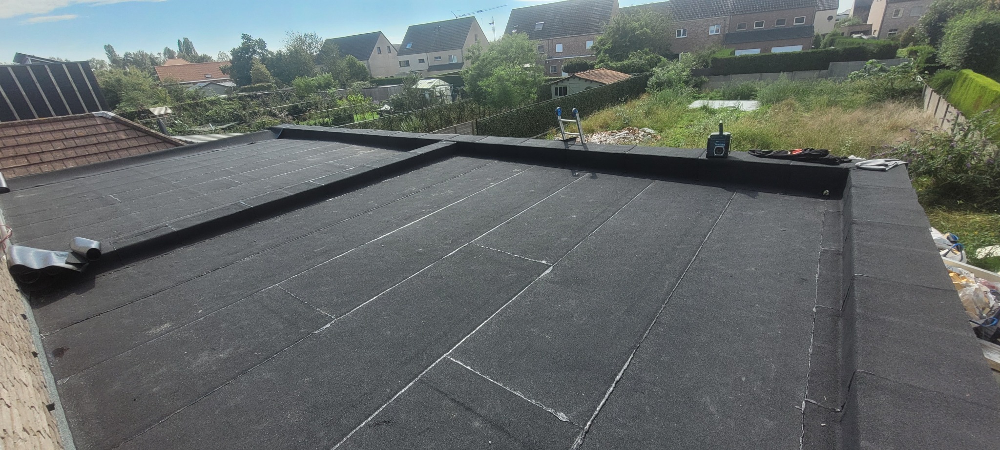 Czarny, płaski dach pokryty papą bitumiczną, z widocznymi łączeniami i fragmentem rolki papy, na tle zieleni i zabudowań mieszkalnych.