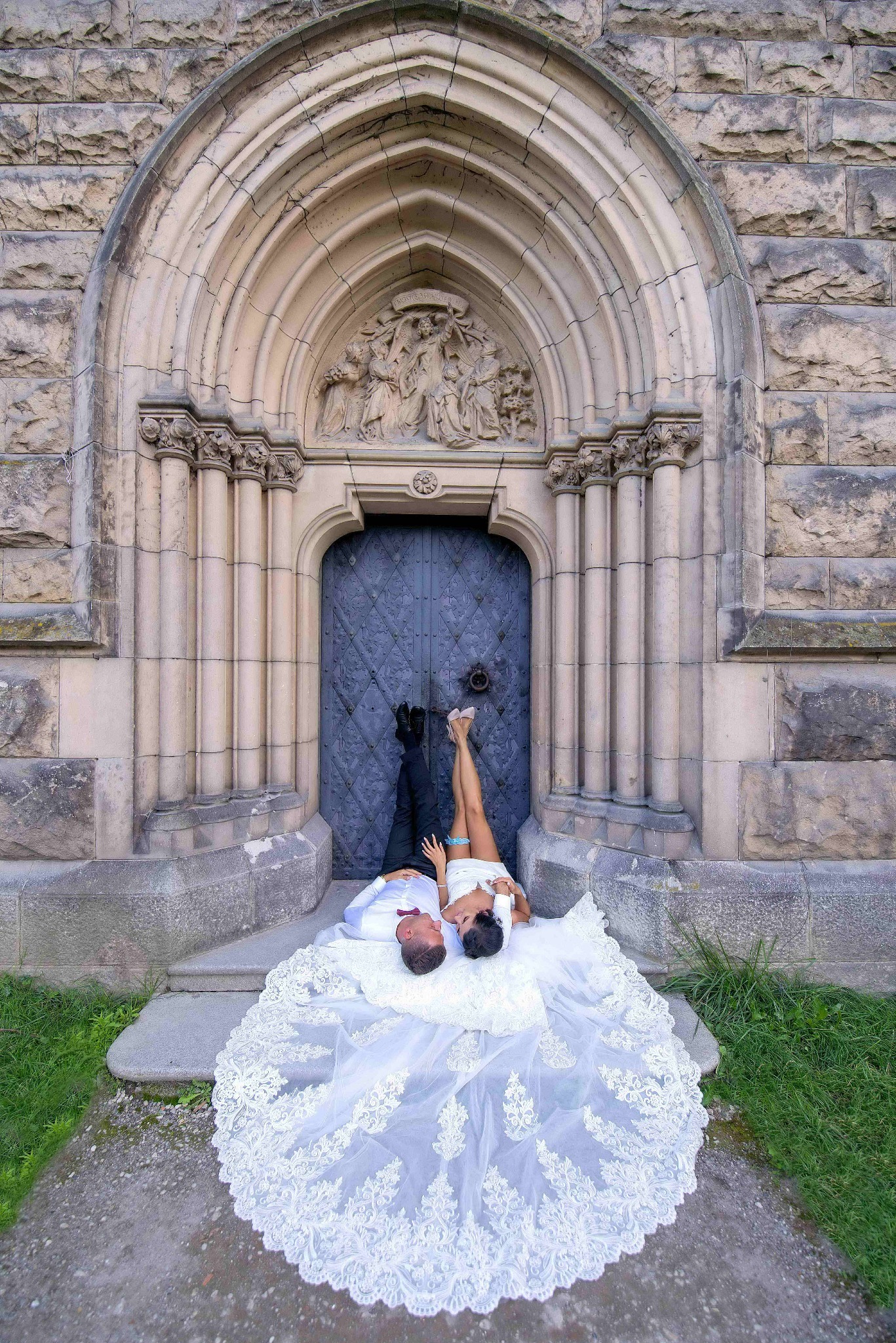 Para młoda leżąca u stóp monumentalnego, kamiennego portalu z rzeźbieniami, suknia ślubna z koronkowym trenem rozłożona na schodach.
