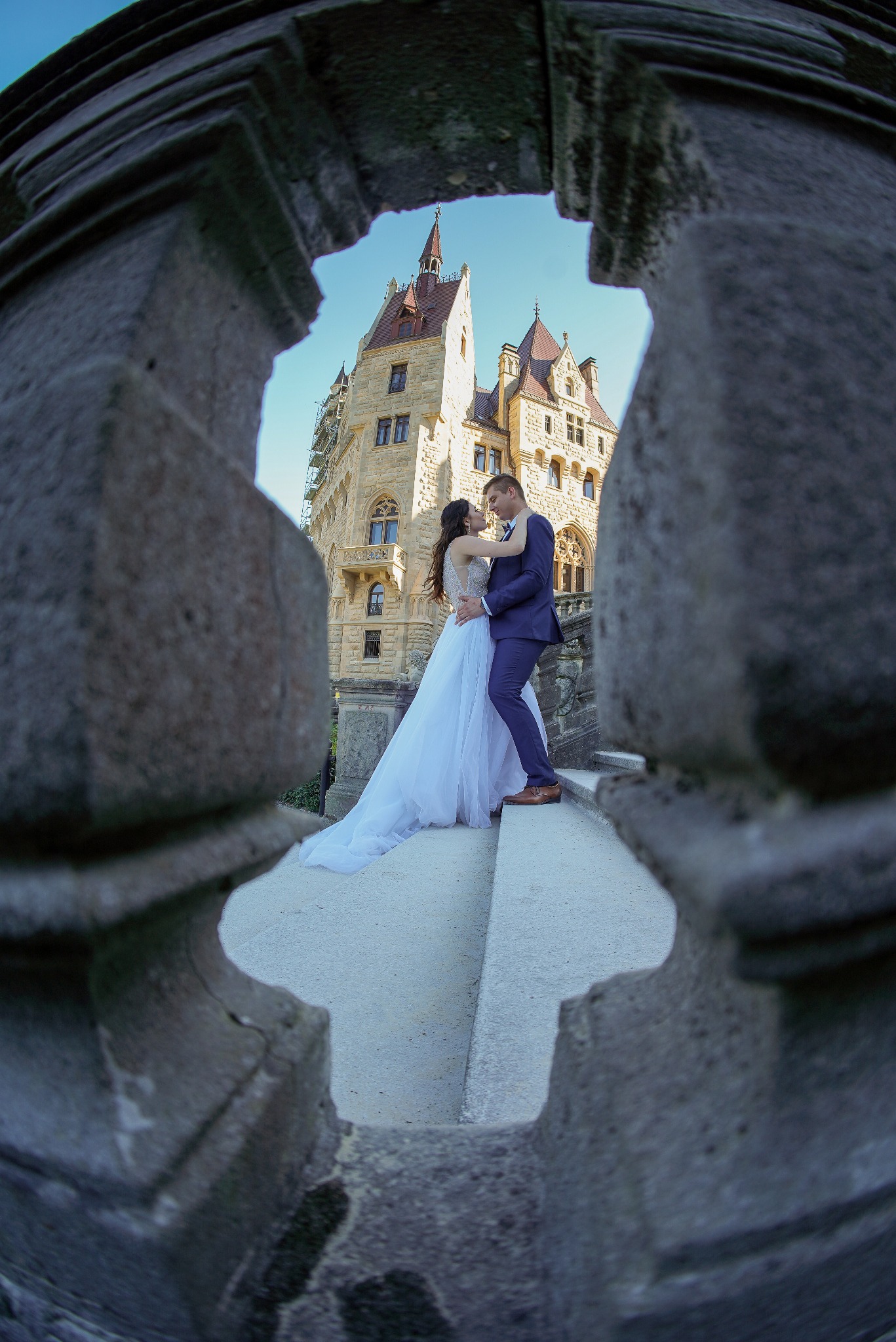 Para młoda w objęciach na tle Zamku Moszna, widok przez kamienną balustradę z dekoracyjnym otworem, suknia ślubna z długim trenem, garnitur w odcieniu granatu.