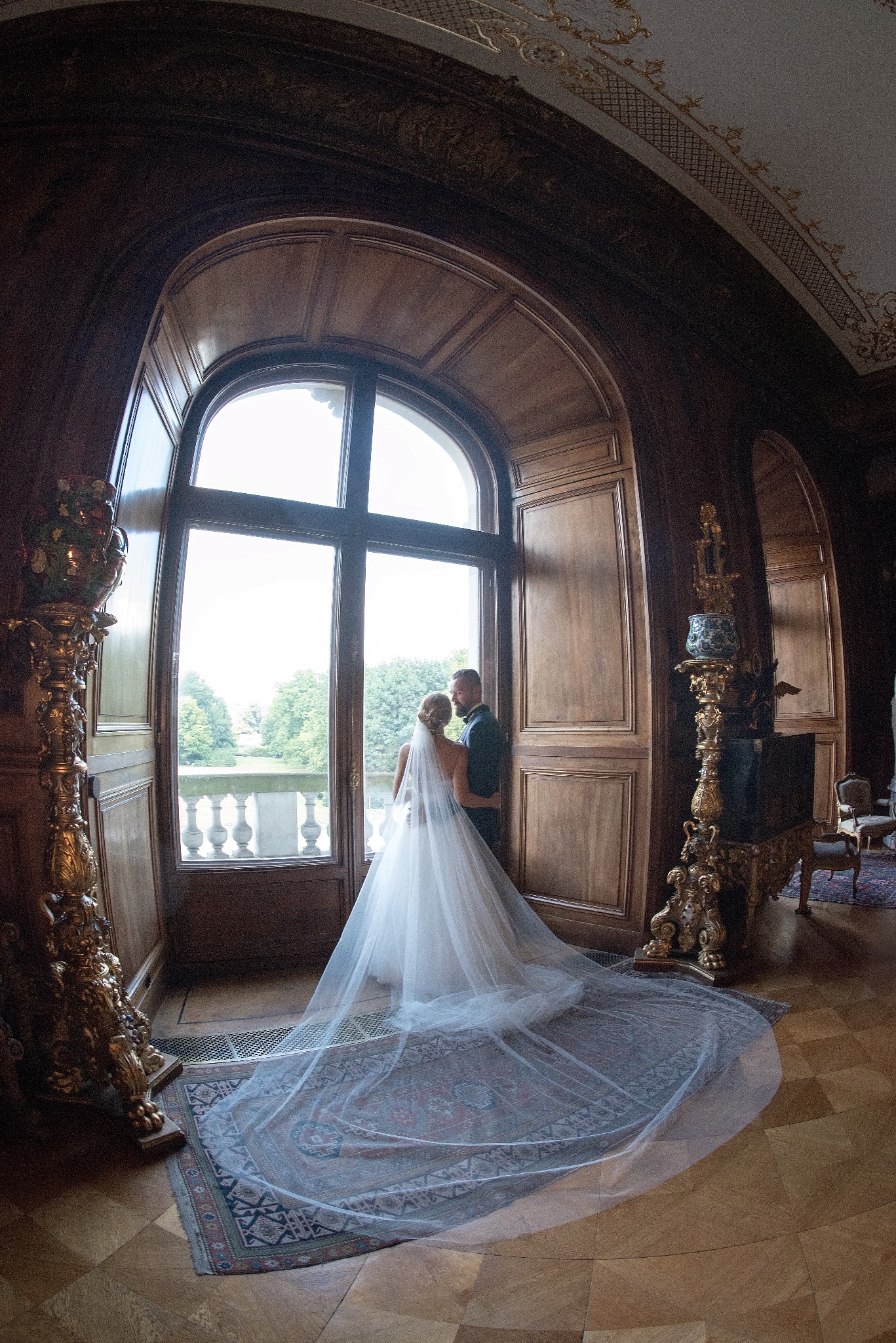 Para młoda w objęciach przy dużym oknie w pałacowym wnętrzu, długi welon rozłożony na dywanie, bogato zdobione kolumny i sufit.