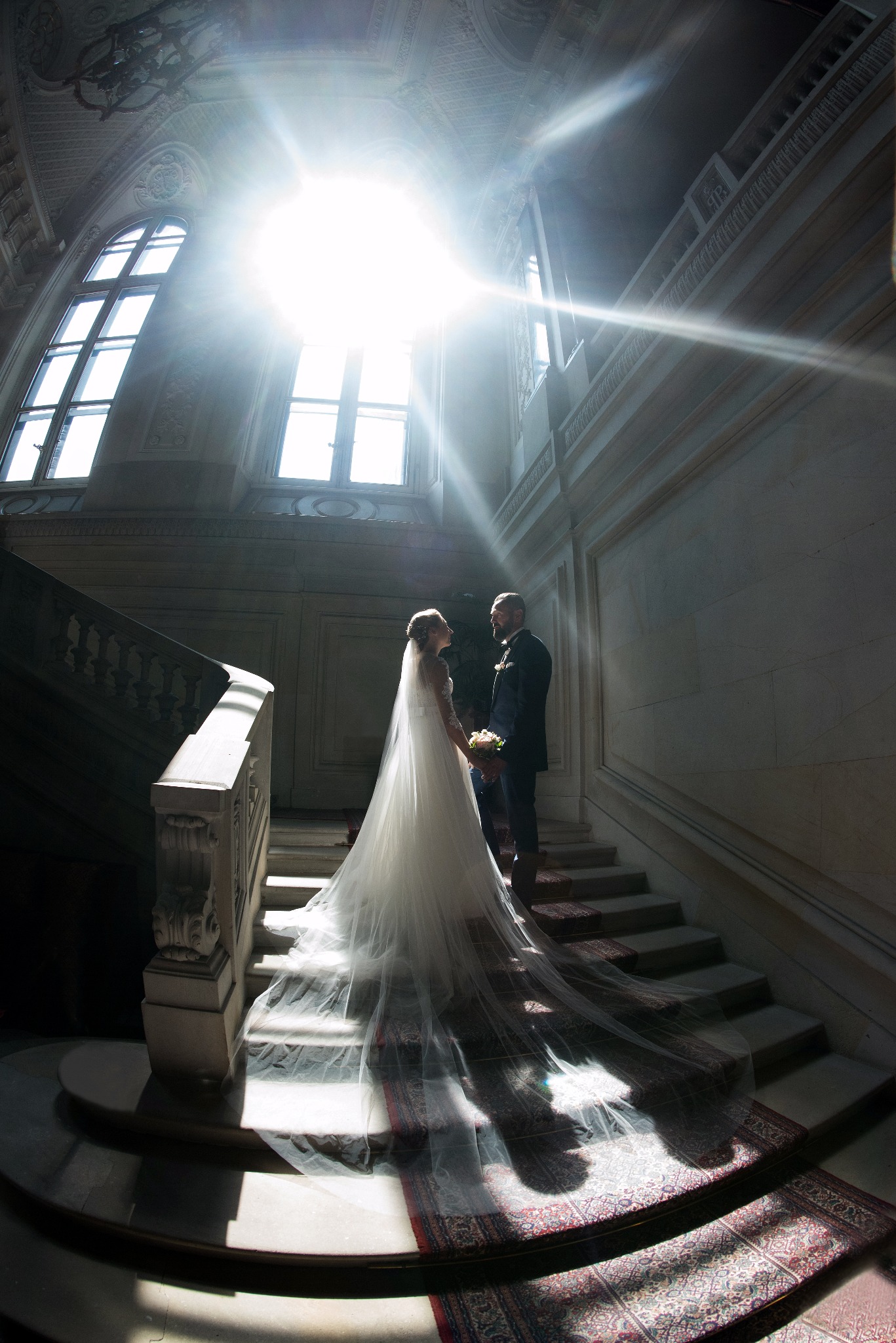Para młoda w Pałacu w Pszczynie, stojąca na schodach w silnym świetle słonecznym wpadającym przez okno, welon panny młodej rozłożony na schodach pokrytych dywanem.