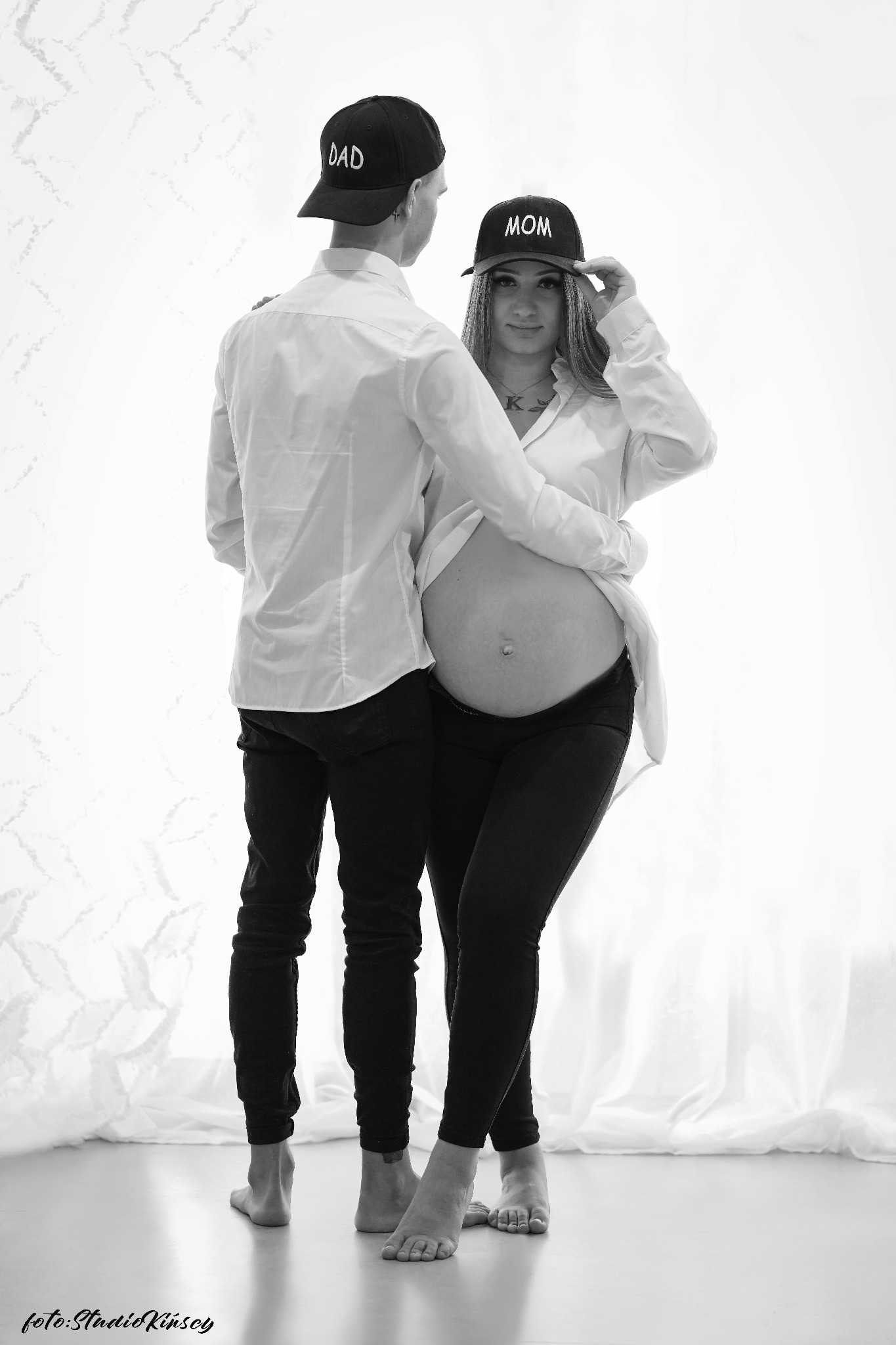 Czarno-białe zdjęcie przyszłych rodziców: kobieta w czapce z napisem 'MOM' i mężczyzna w czapce z napisem 'DAD', obejmujący ciążowy brzuch, na jasnym tle.