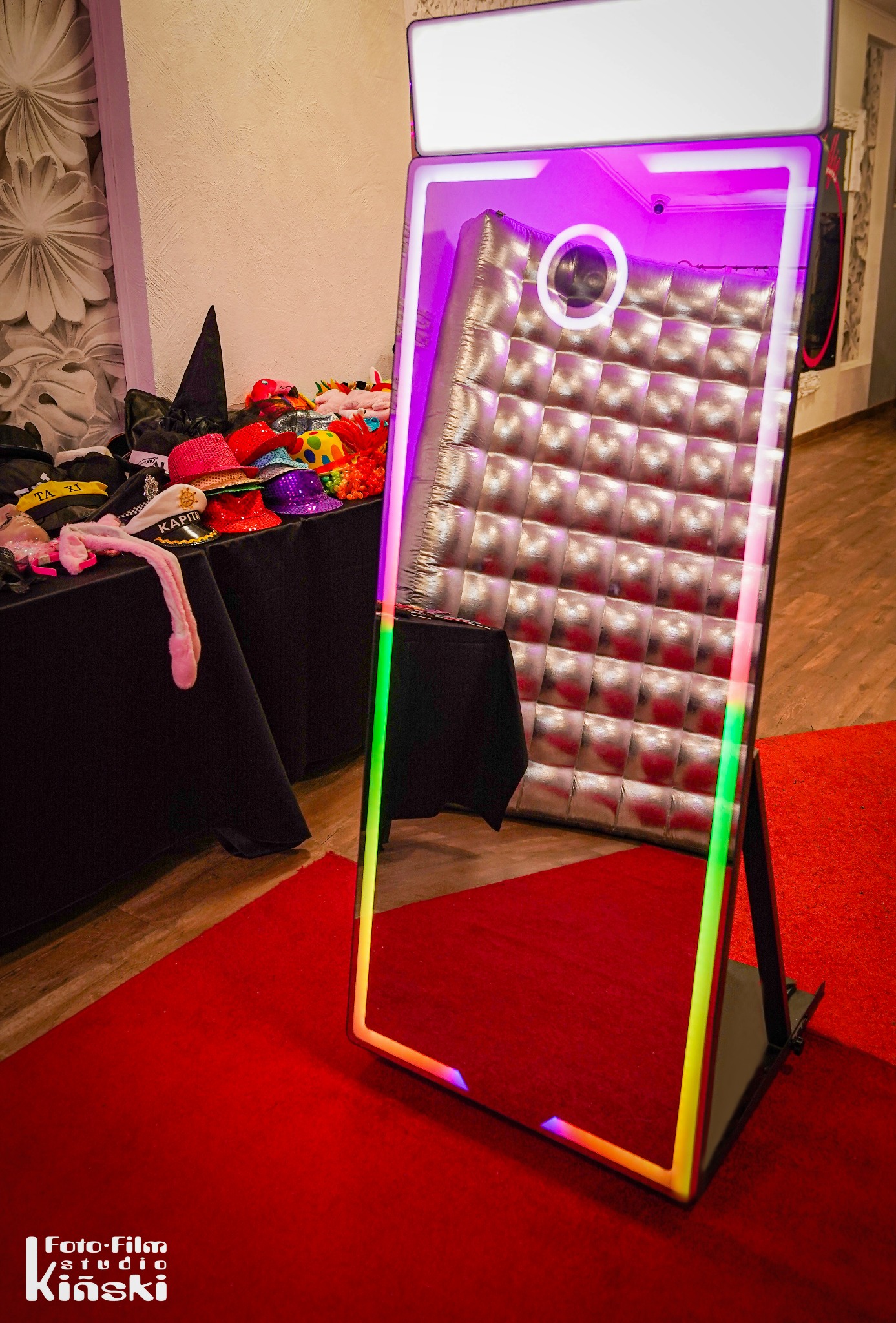 Foto-lustro z kolorowym oświetleniem na czerwonym dywanie, obok stolik z rekwizytami (kapelusze, peruki). Tło stanowi ściana z kwiatowym wzorem.