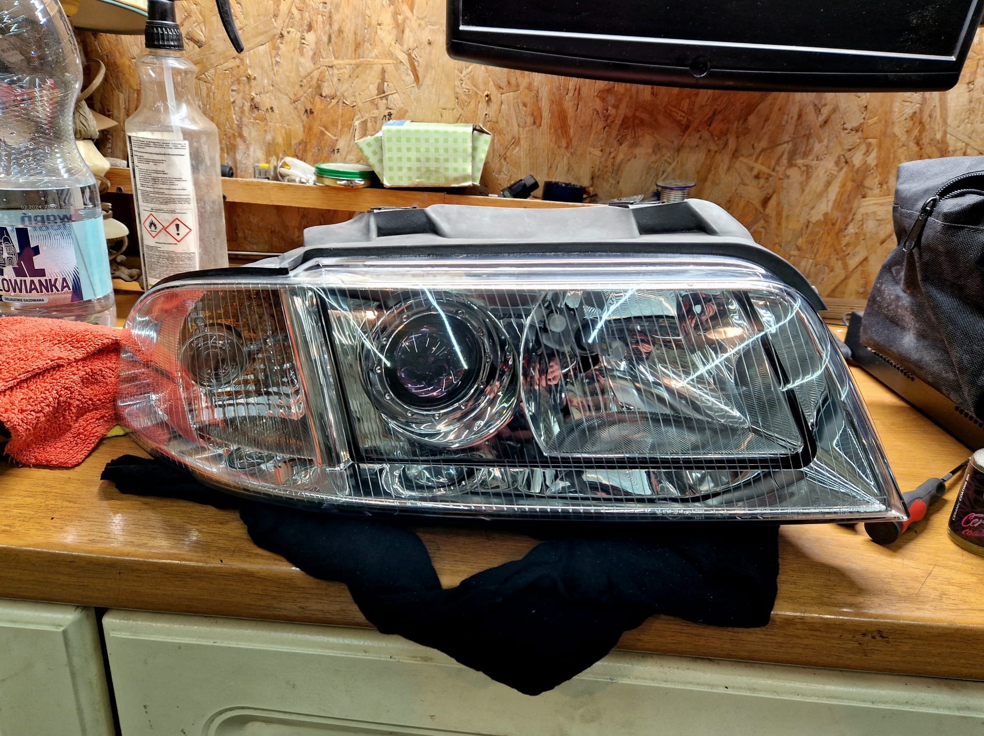 Wyczyszczona lampa samochodowa na warsztatowym stole, widoczne refleksy światła, butelki z płynami i narzędzia, co sugeruje etap detailingu lub renowacji.