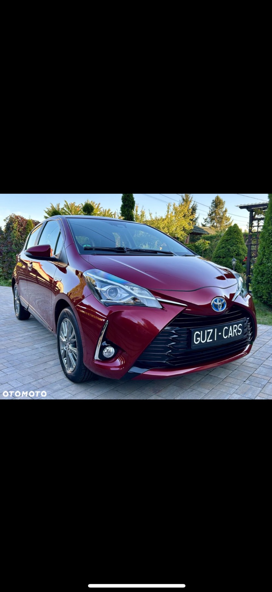 Bordowy Toyota Yaris po myciu, z widocznym logo firmy detailingowej na tablicy rejestracyjnej, zaparkowany na kostce brukowej w ogrodzie.
