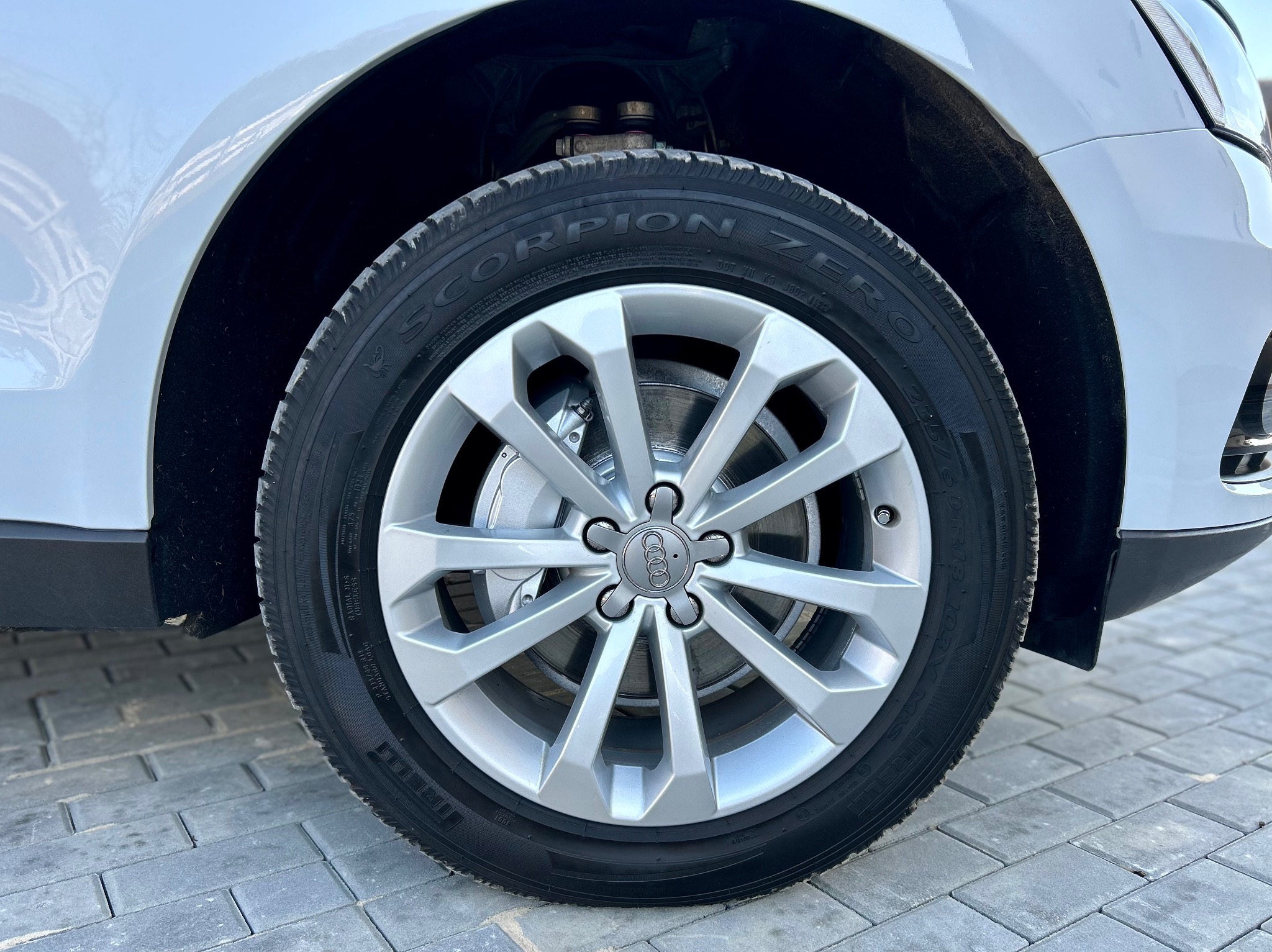 Błyszczące, srebrne felgi i opony Pirelli Scorpion Zero na białym Audi Q5, zaparkowanym na szarej kostce brukowej. Widok z bliska na przednie koło.