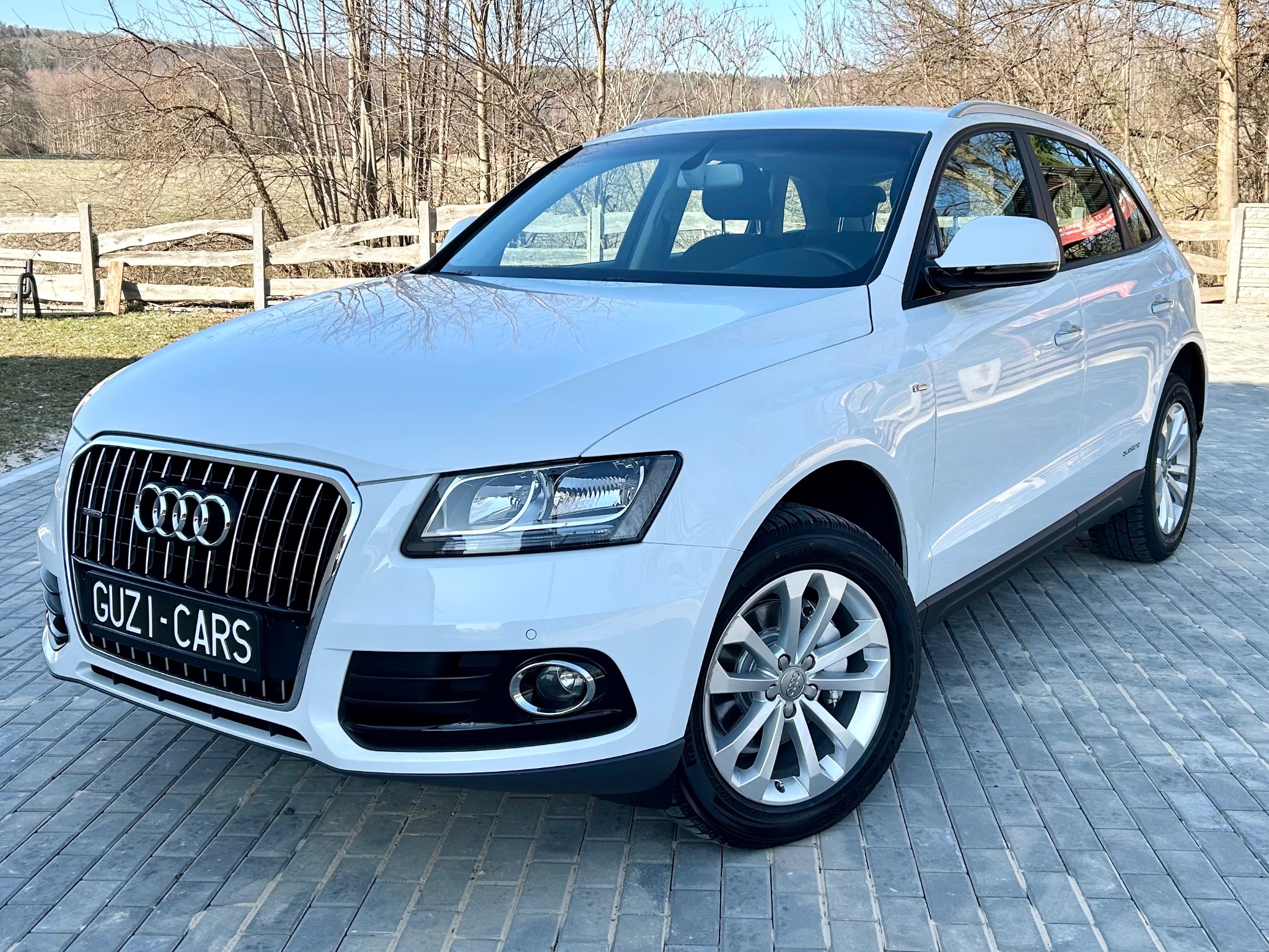 Białe Audi Q5 po usłudze mycia, zaparkowane na kostce brukowej, z widocznym logo firmy na tablicy rejestracyjnej i elementami krajobrazu w tle.