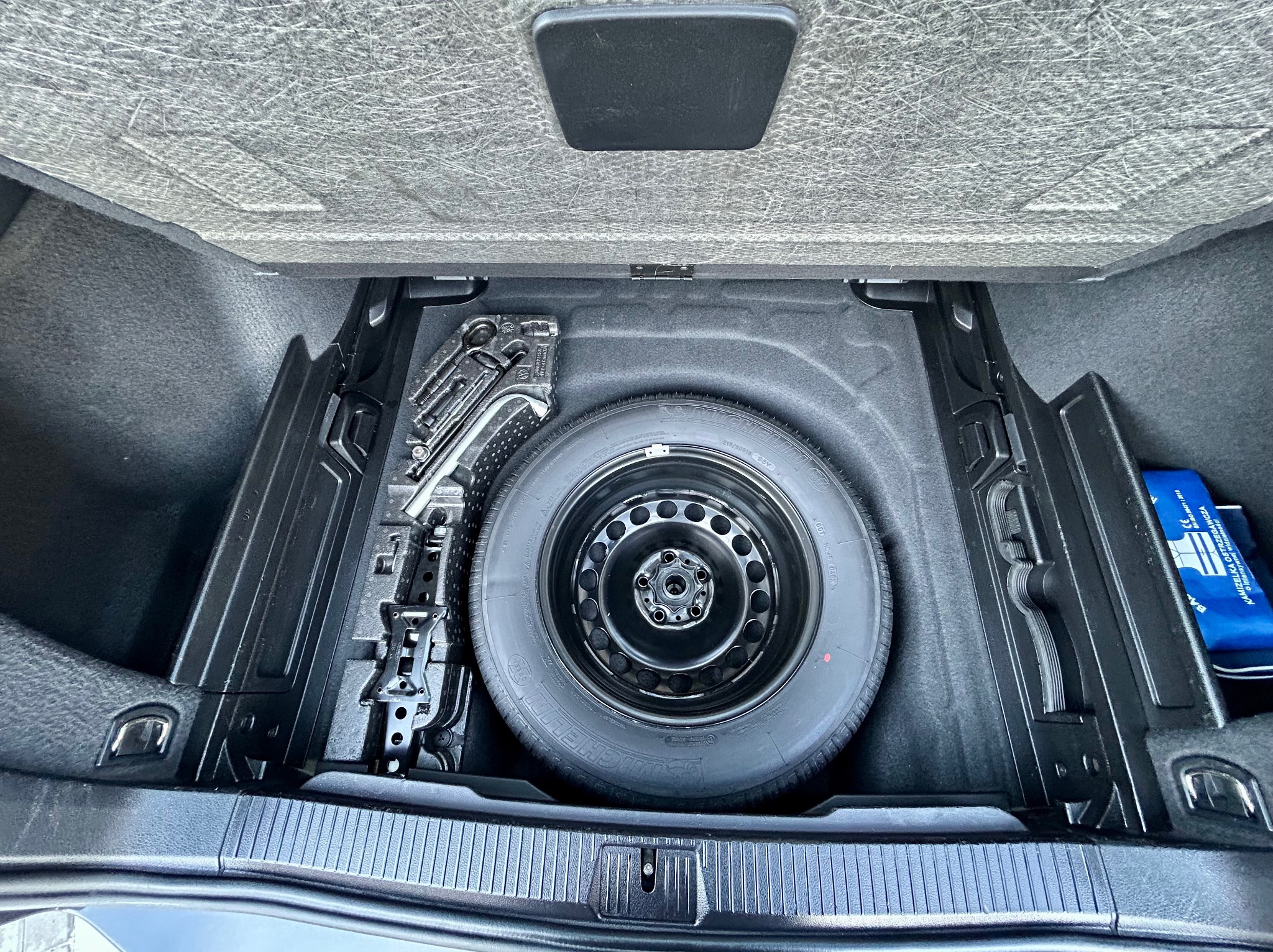 Wnętrze bagażnika samochodu z kołem zapasowym, zestawem narzędzi i apteczką. Widok z góry, oświetlenie naturalne, czarne elementy wykończenia.