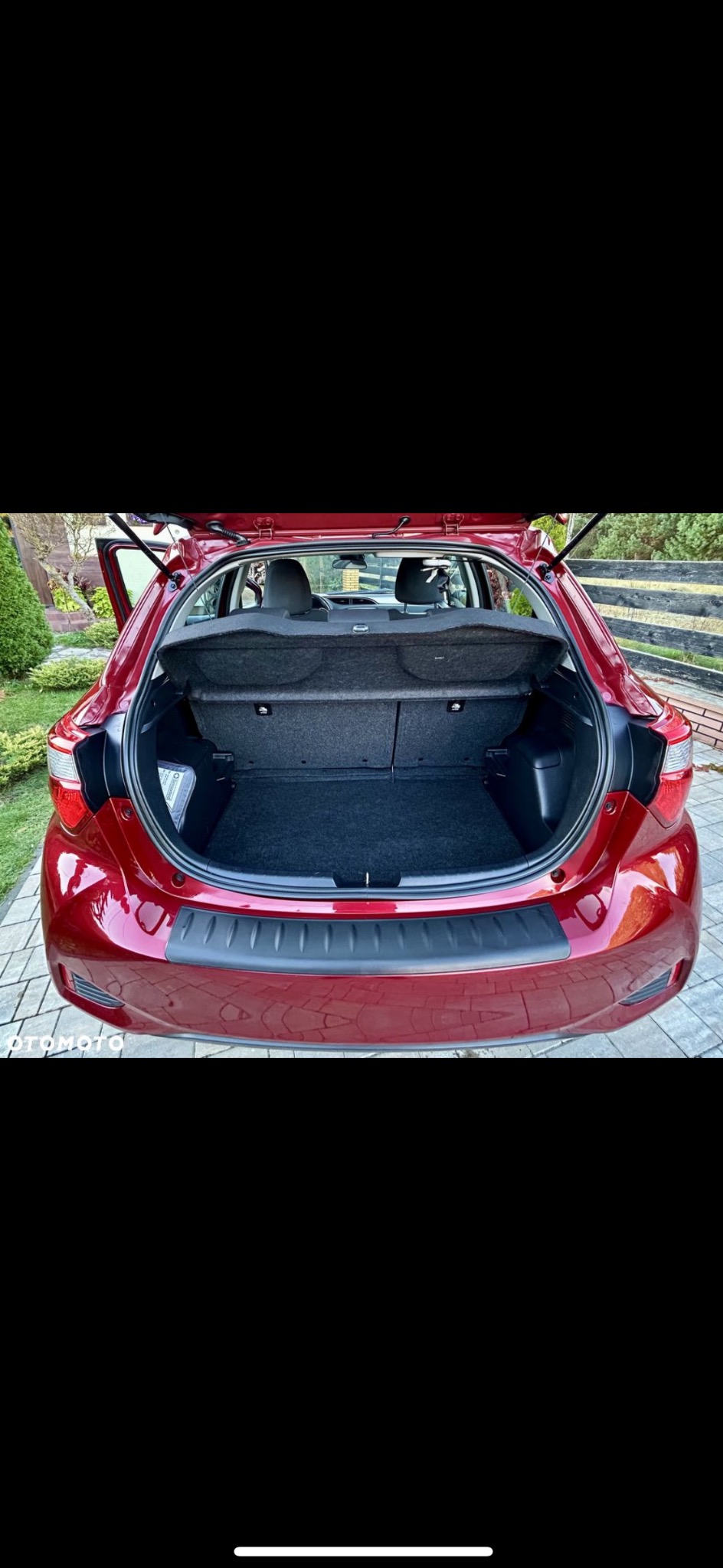 Czerwony samochód z otwartym bagażnikiem, widok z tyłu. Czyste wnętrze bagażnika, zadbany lakier. Otoczenie to trawnik i kostka brukowa.