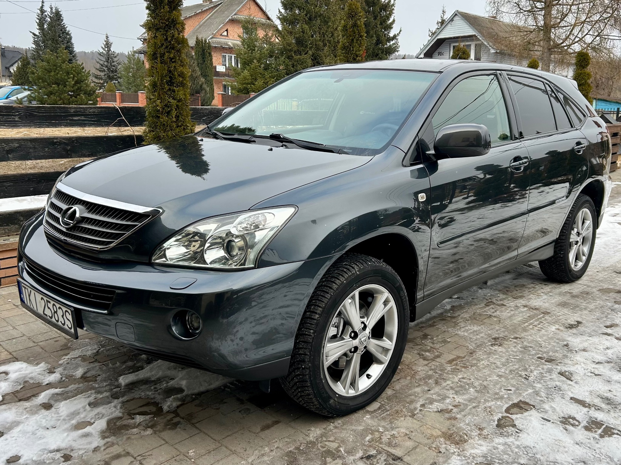 Szary SUV Lexus na kostce brukowej z resztkami śniegu, na tle domów i drzew. Widok z przodu, lekko z boku. Tablica rejestracyjna widoczna.