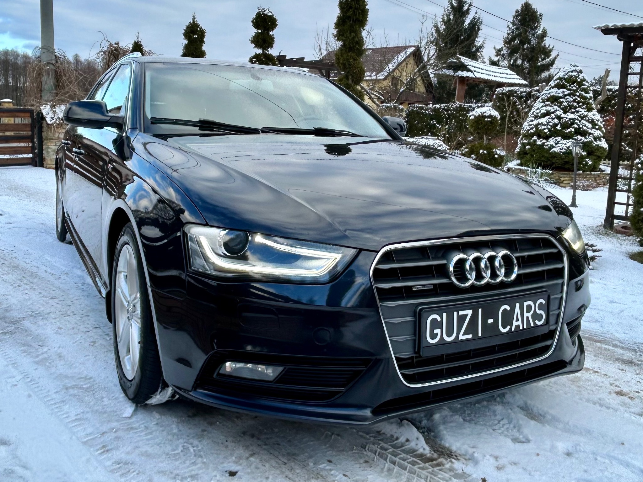 Błyszczący, czarny samochód Audi, zaparkowany na śniegu, z widocznym logo i tablicą 'GUZI-CARS'. W tle dom i dekoracyjne drzewka.