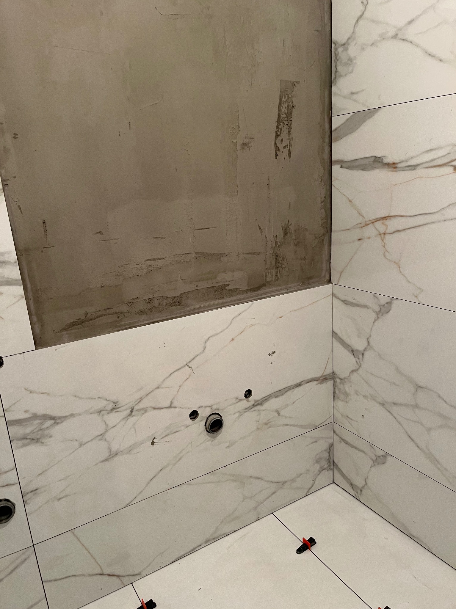 Łazienka w trakcie wykańczania: białe płytki imitujące marmur, częściowo położone na ścianie i podłodze. Widoczne otwory na instalacje i czerwone kliny glazurnicze.