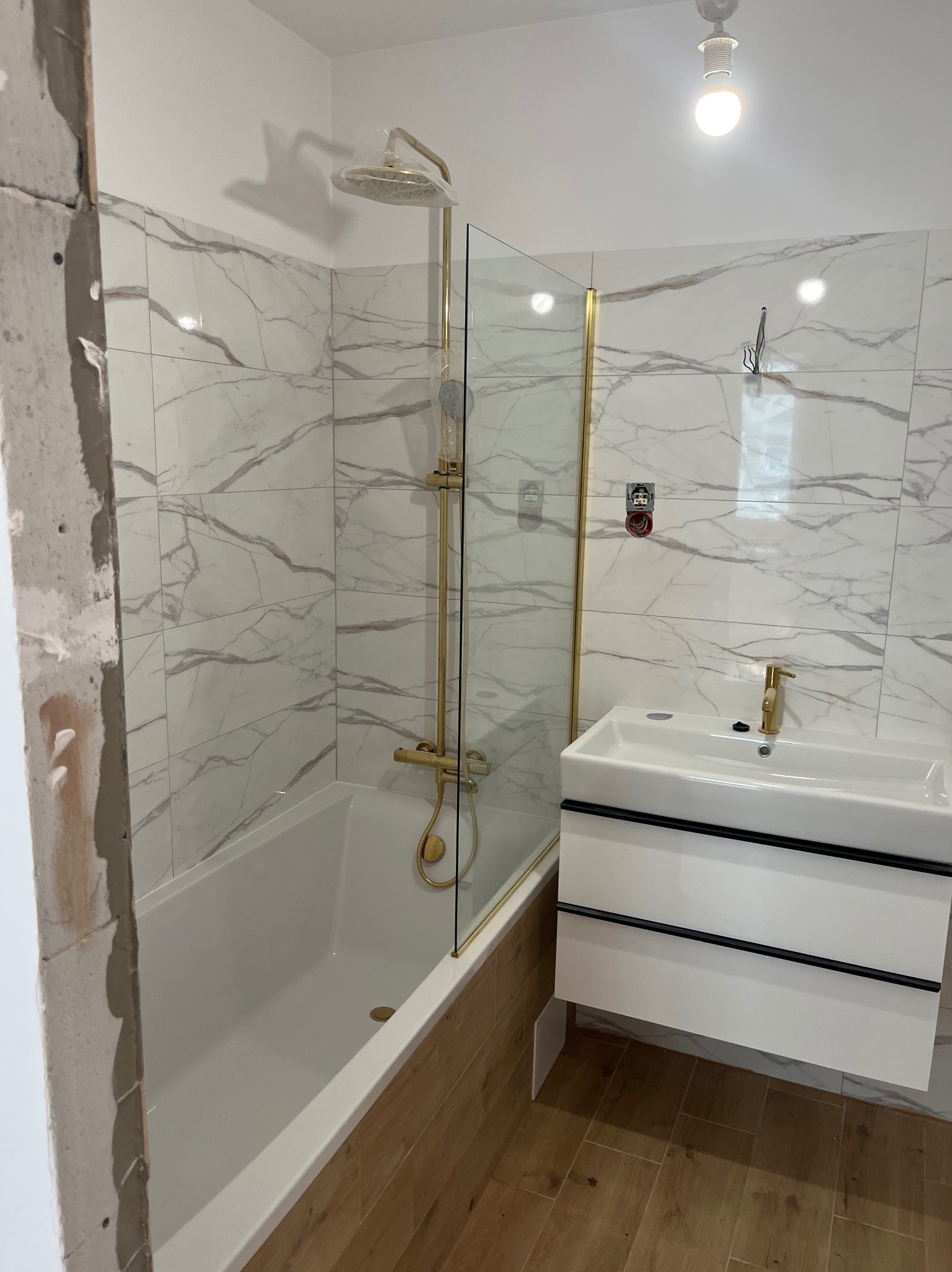Nowoczesna łazienka w trakcie wykańczania: białe płytki imitujące marmur, złota armatura, biała umywalka z szafką i fragment surowej ściany.