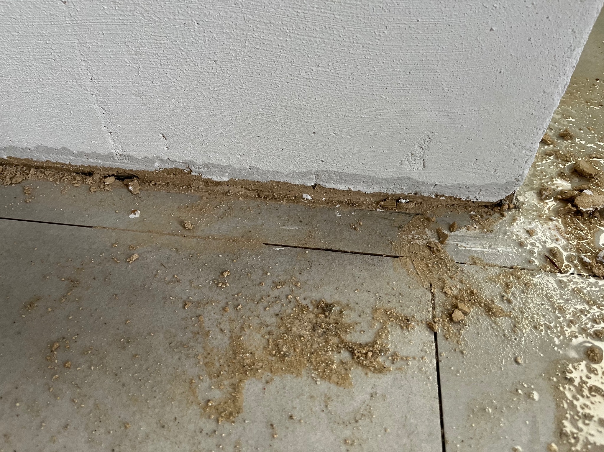Narożnik ściany i podłogi w trakcie prac wykończeniowych. Widoczne zabrudzenia i resztki materiałów budowlanych na szarej, błyszczącej podłodze.