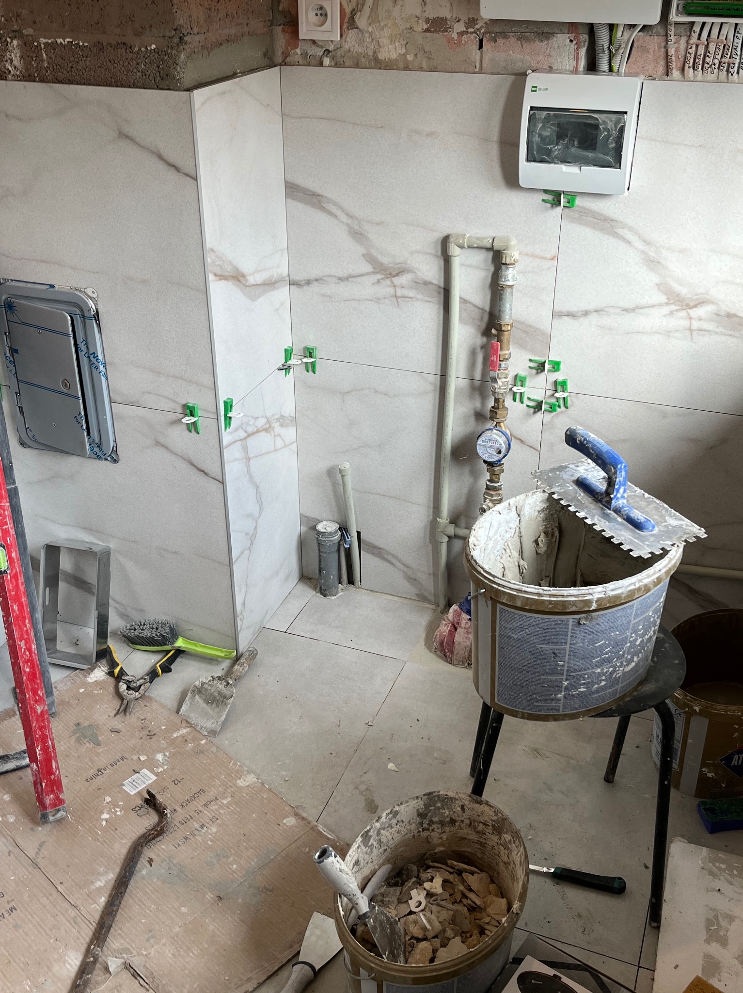 Łazienka w trakcie prac glazurniczych: marmurowe płytki, poziomica, wiadro z zaprawą, krzyżyki dystansowe, widoczne rury i skrzynka elektryczna.
