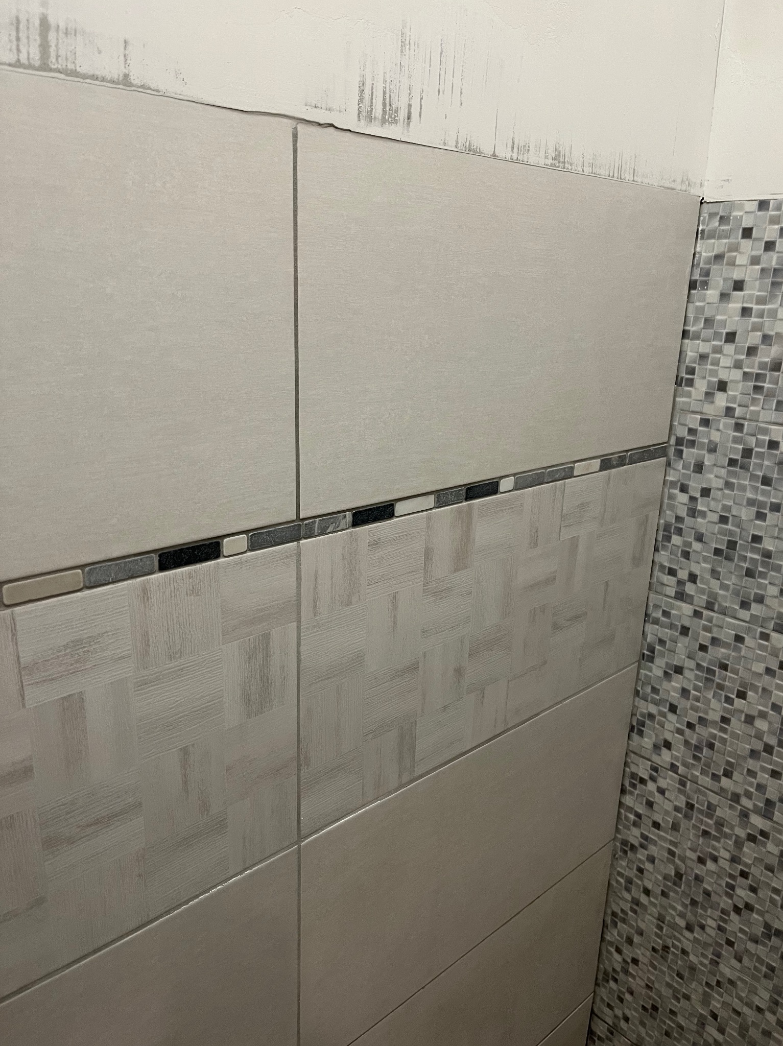 Ściana łazienki w trakcie wykańczania w Warszawie: ułożone płytki ceramiczne w odcieniach szarości, z dekoracyjnym pasem i mozaiką po prawej stronie. Widoczne niedociągnięcia.