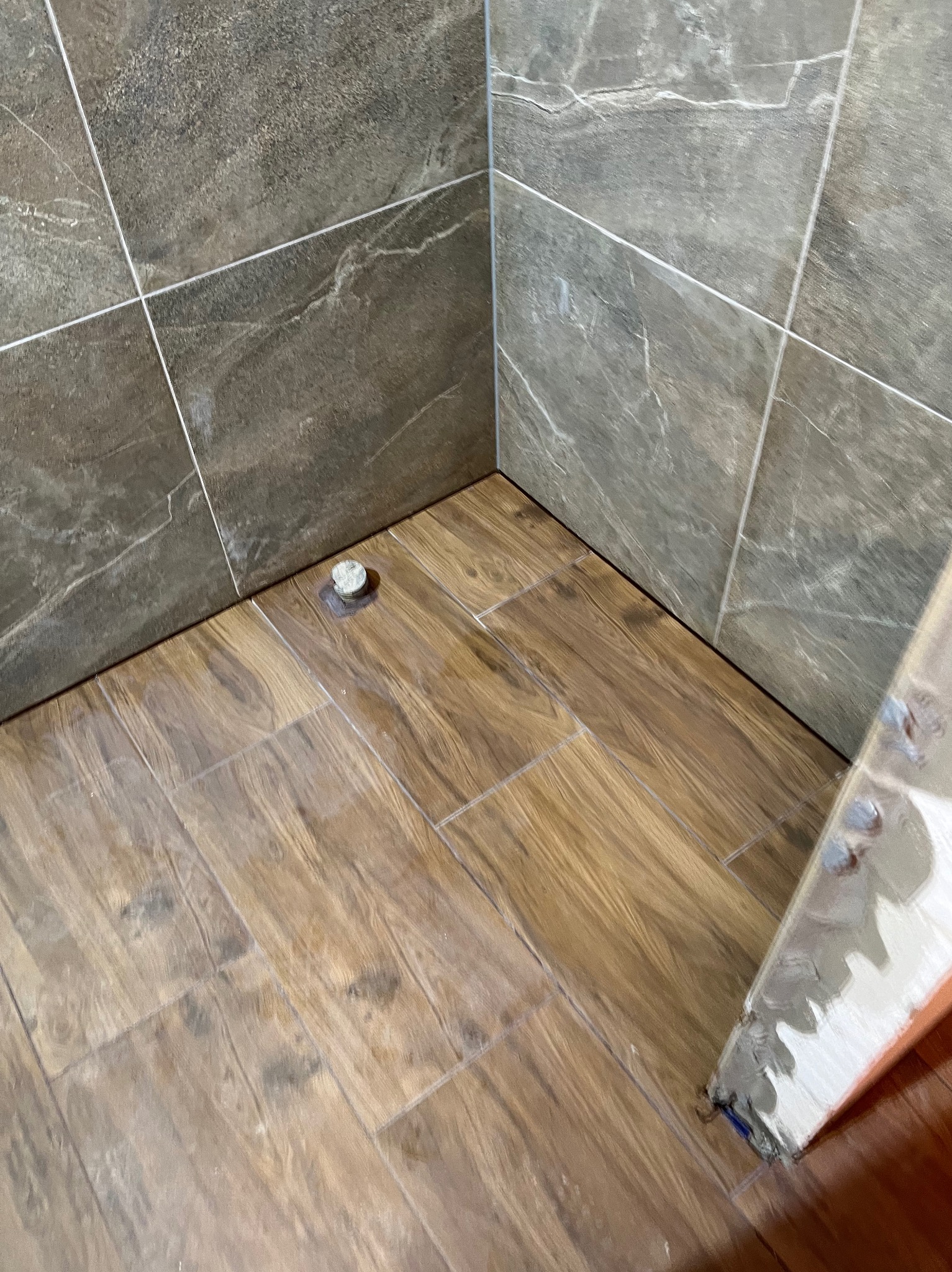 Ukończona podłoga i fragment ściany w rogu łazienki, wyłożone płytkami imitującymi drewno i marmur. Widoczna fuga i narzędzie glazurnicze.