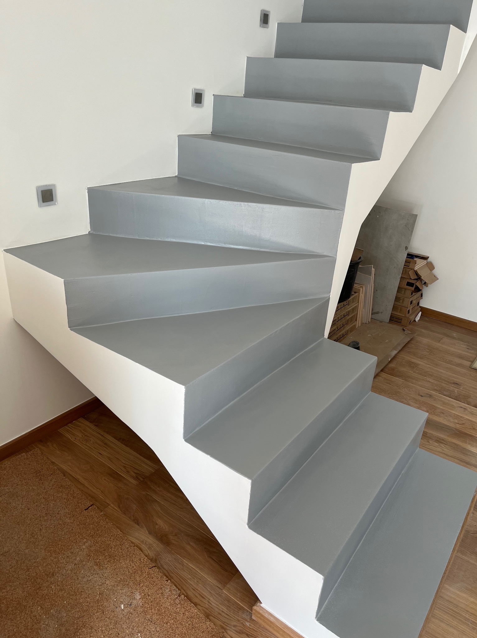 Nowoczesne, betonowe schody pomalowane na szaro, z białymi bokami, oświetlenie punktowe na ścianie. Widoczne drewniane panele podłogowe.