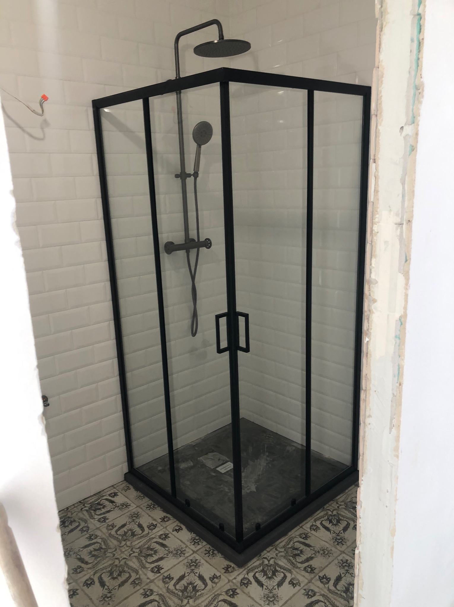 Nowoczesna kabina prysznicowa z czarną ramą i deszczownicą, wykończona białymi płytkami imitującymi cegłę i wzorzystą podłogą w łazience w trakcie remontu.