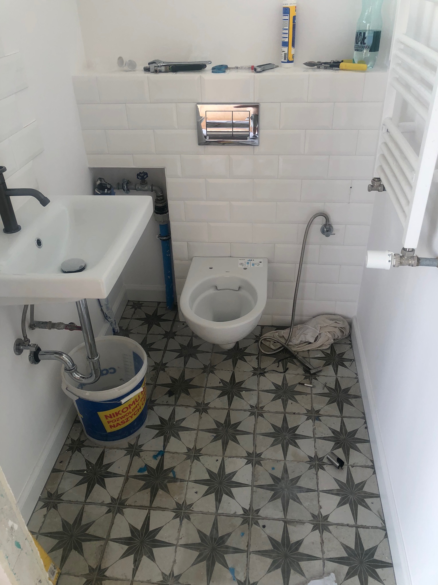 Biała łazienka w trakcie montażu armatury: umywalka z czarną baterią, wisząca toaleta, grzejnik drabinkowy. Na podłodze wzorzyste płytki. Widoczne narzędzia i wiadro.