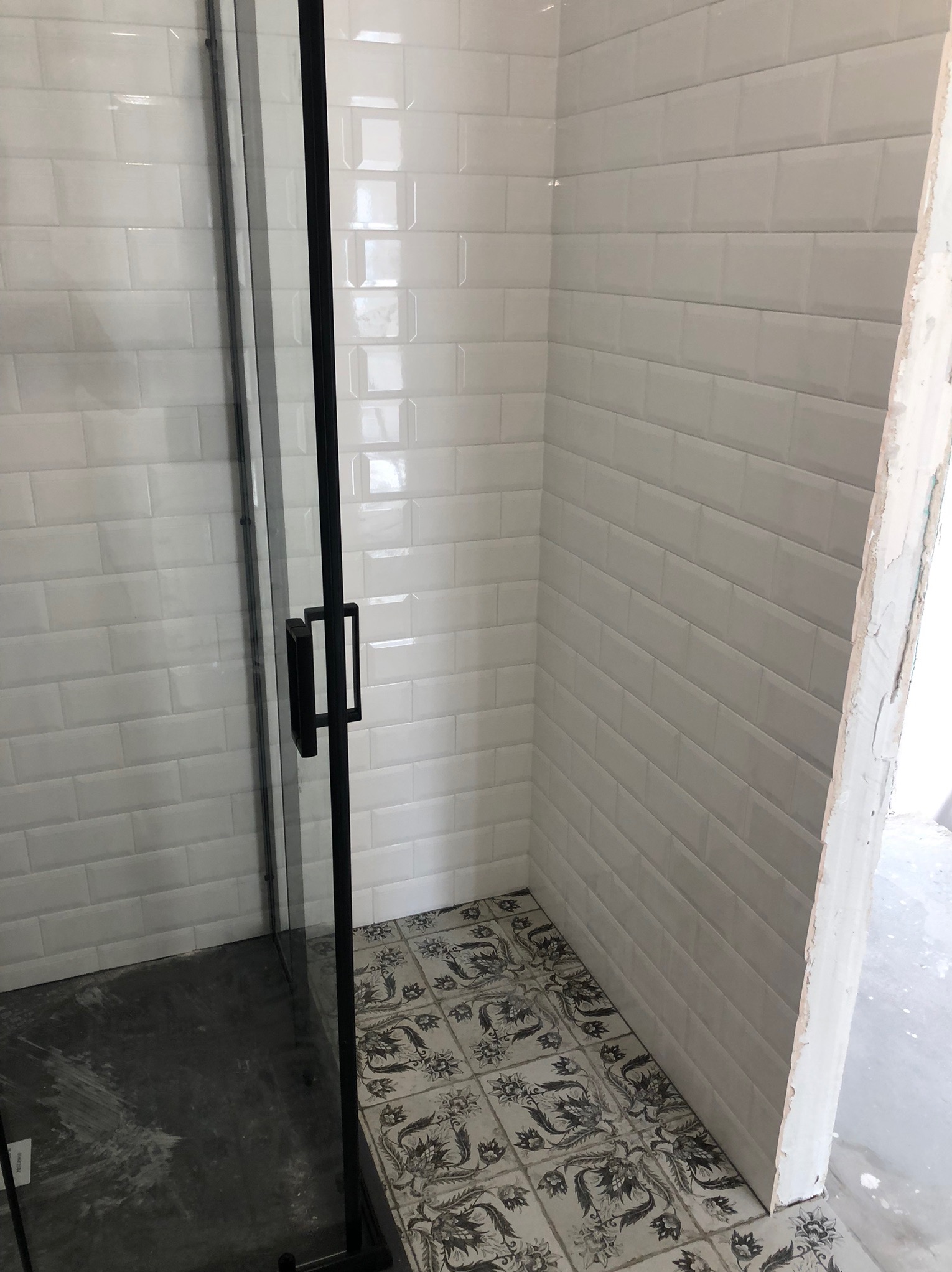 Wykończona kabina prysznicowa z czarnymi detalami, białe kafelki na ścianach ułożone w cegiełkę, podłoga z wzorzystych płytek. Widok na fragment łazienki w trakcie prac wykończeniowych.