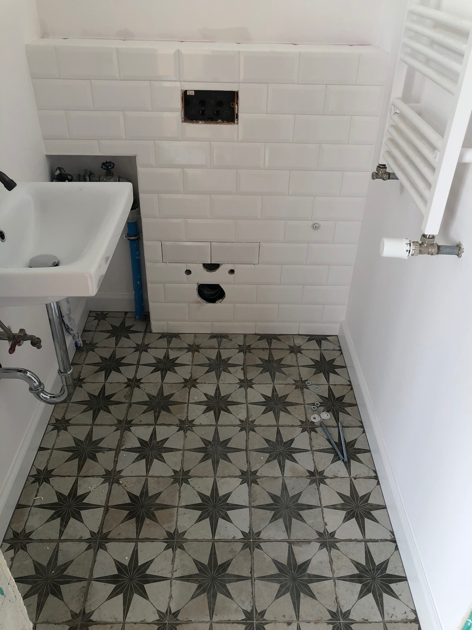 Łazienka w trakcie remontu: białe płytki na ścianach, wzorzysta podłoga, umywalka i grzejnik. Widoczne otwory instalacyjne i narzędzia.