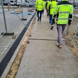 Bud-Safe BHP OŚ - Grupa pracowników w odblaskowych kurtkach i kaskach idzie chodnikiem obok metalowej kratki odwadniającej na placu budowy z kontenerami biurowymi w tle.