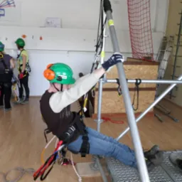 Uczestnik w zielonym kasku i uprzęży, demonstruje technikę asekuracji na rusztowaniu podczas zajęć praktycznych w sali szkoleniowej, w tle dwie osoby obserwują ćwiczenia.