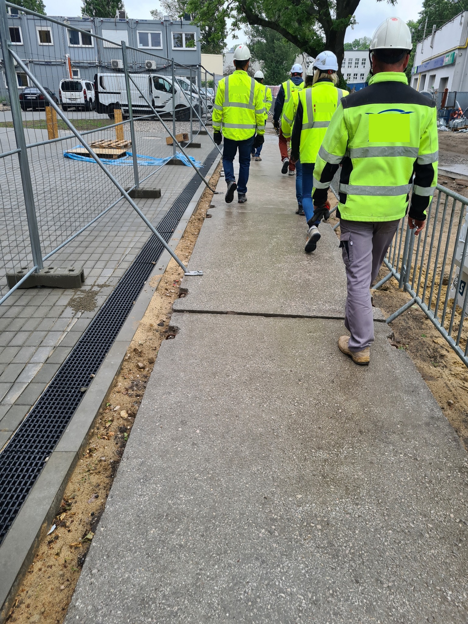 Grupa pracowników w odblaskowych kurtkach i kaskach idzie chodnikiem obok metalowej kratki odwadniającej na placu budowy z kontenerami biurowymi w tle.