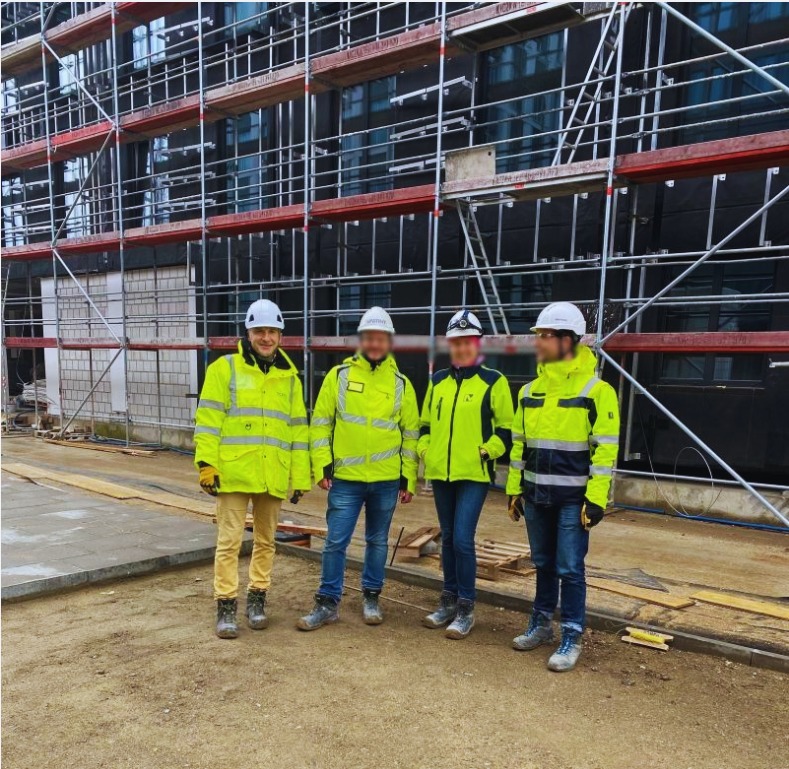 Grupa czterech osób w odblaskowych kurtkach i kaskach ochronnych stoi na placu budowy przed budynkiem w rusztowaniach. Jeden mężczyzna uśmiecha się, pozostałe twarze są zamazane.