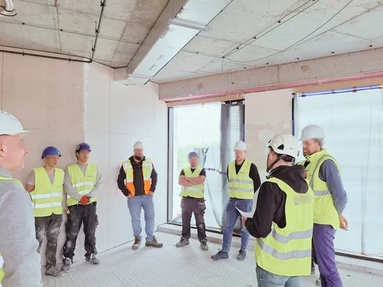 Grupa osób w kaskach i kamizelkach odblaskowych, prawdopodobnie uczestniczących w szkoleniu BHP na placu budowy w surowym stanie, z widocznymi instalacjami pod sufitem.
