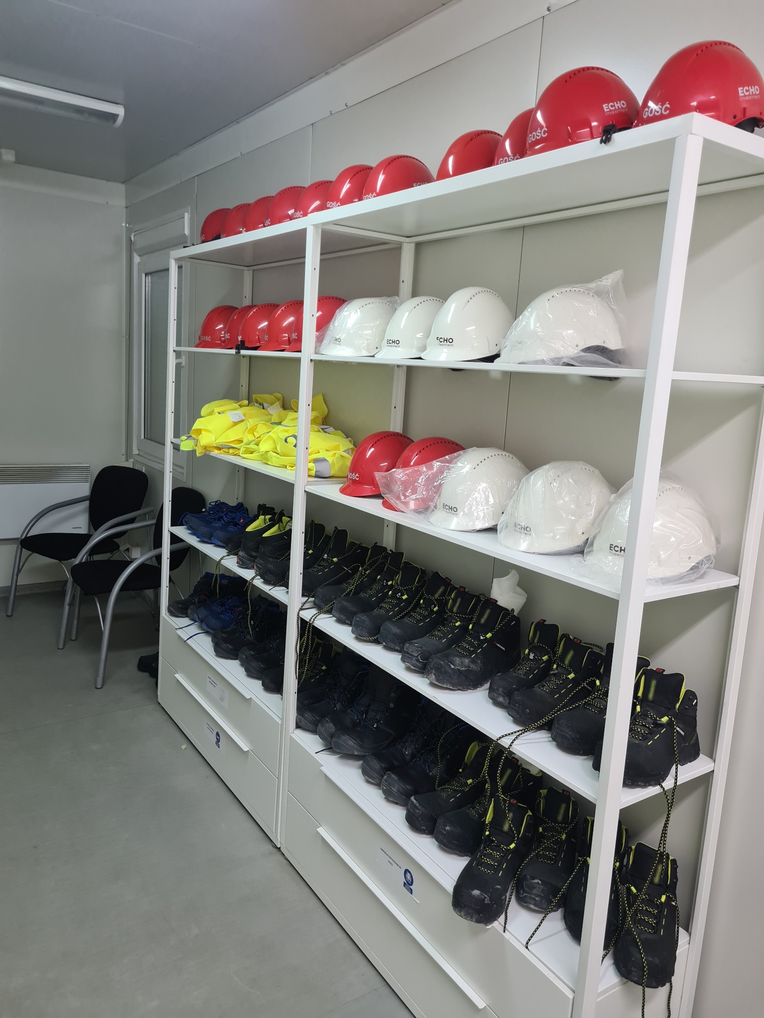 Regał z odzieżą i obuwiem roboczym: czerwone i białe kaski z logo, żółte kamizelki odblaskowe i czarne buty robocze z żółtymi sznurówkami, ułożone na półkach w pomieszczeniu socjalnym.