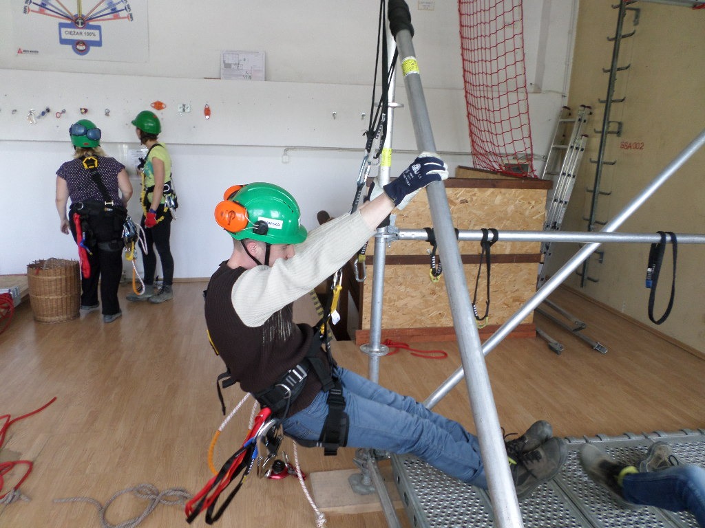 Uczestnik w zielonym kasku i uprzęży, demonstruje technikę asekuracji na rusztowaniu podczas zajęć praktycznych w sali szkoleniowej, w tle dwie osoby obserwują ćwiczenia.
