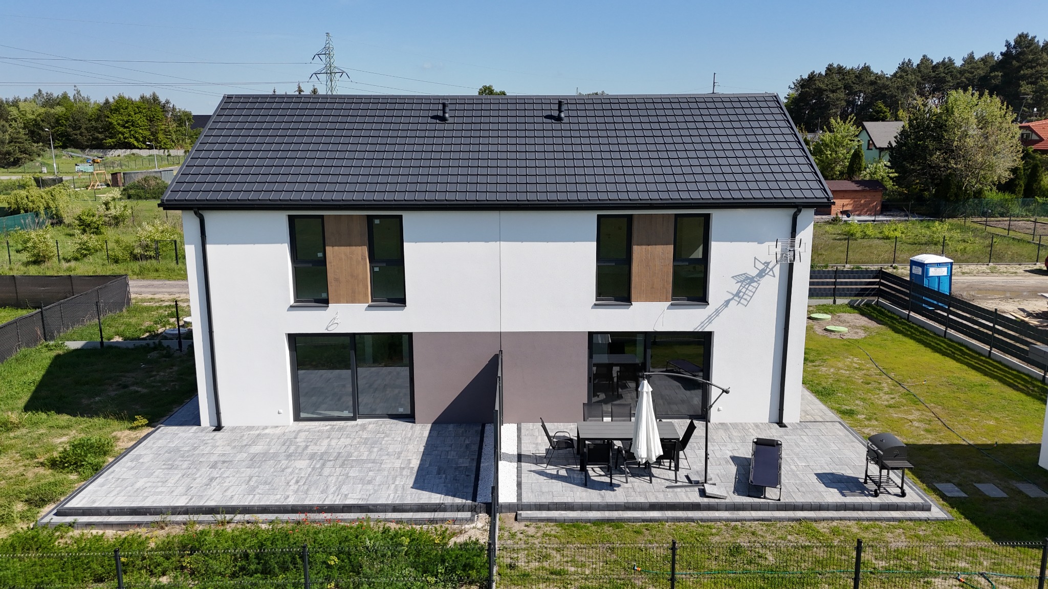 Nowoczesny, dwukondygnacyjny dom bliźniak z szarym dachem, białymi ścianami i dużymi oknami. Taras z meblami ogrodowymi i grillem. Widok z góry, słoneczny dzień.