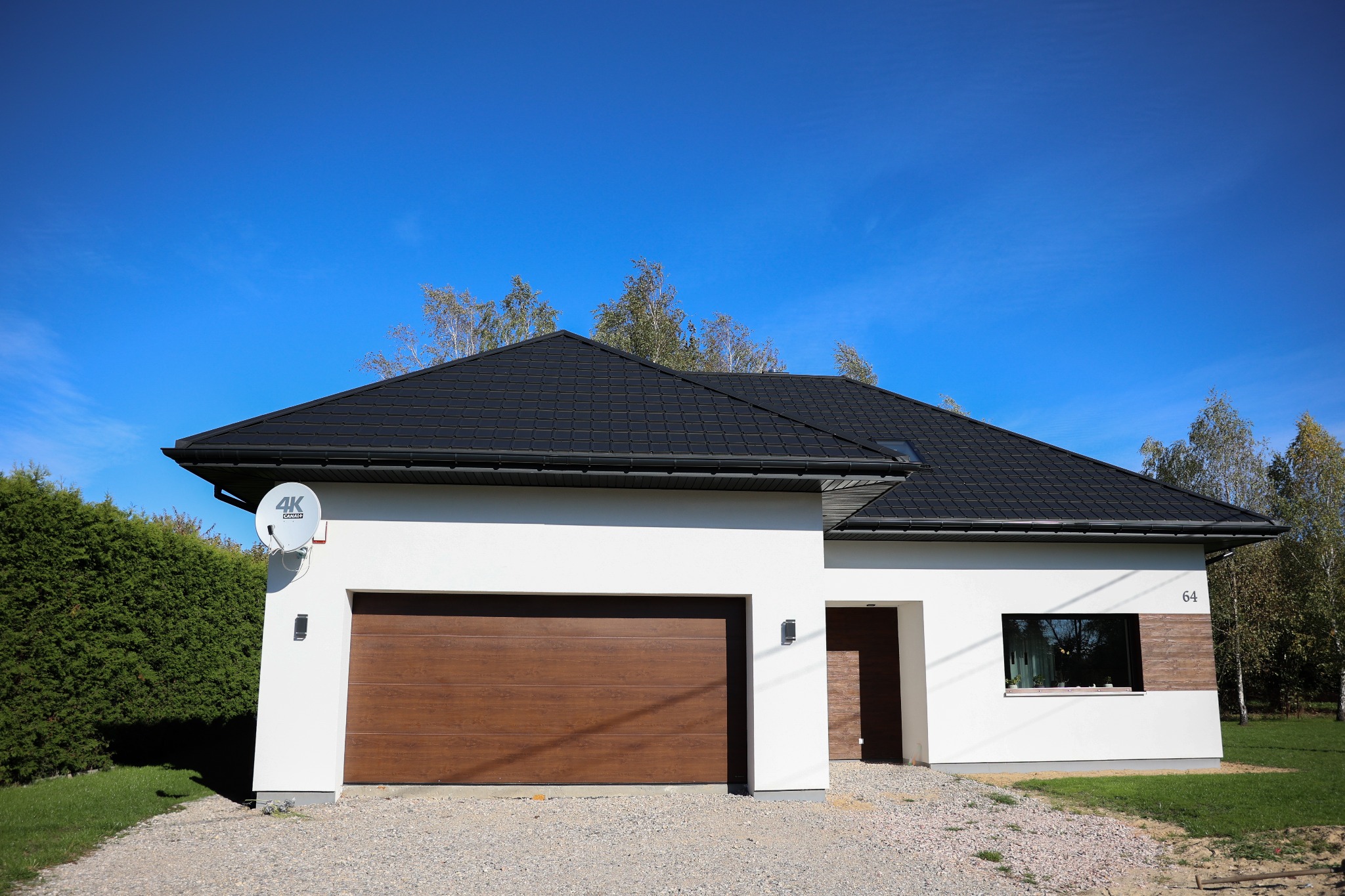 Nowoczesny dom z białą elewacją, brązową bramą garażową i dachem z czarnej dachówki. Widoczna antena satelitarna i numer domu '64'.