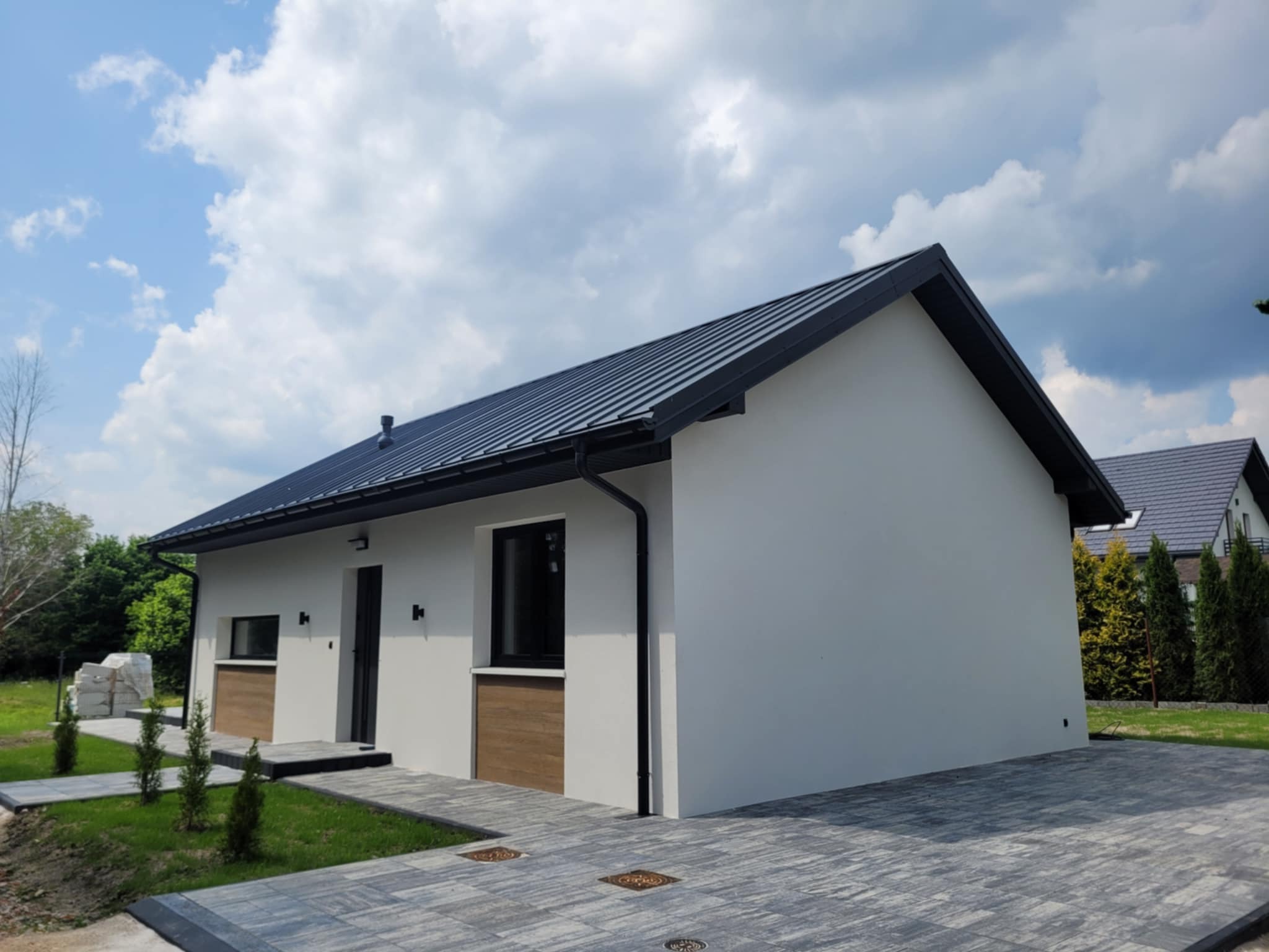 Nowoczesny, parterowy dom z ciemnym dachem i szarą kostką brukową na podjeździe w słoneczny dzień. Widoczne elementy wykończeniowe.