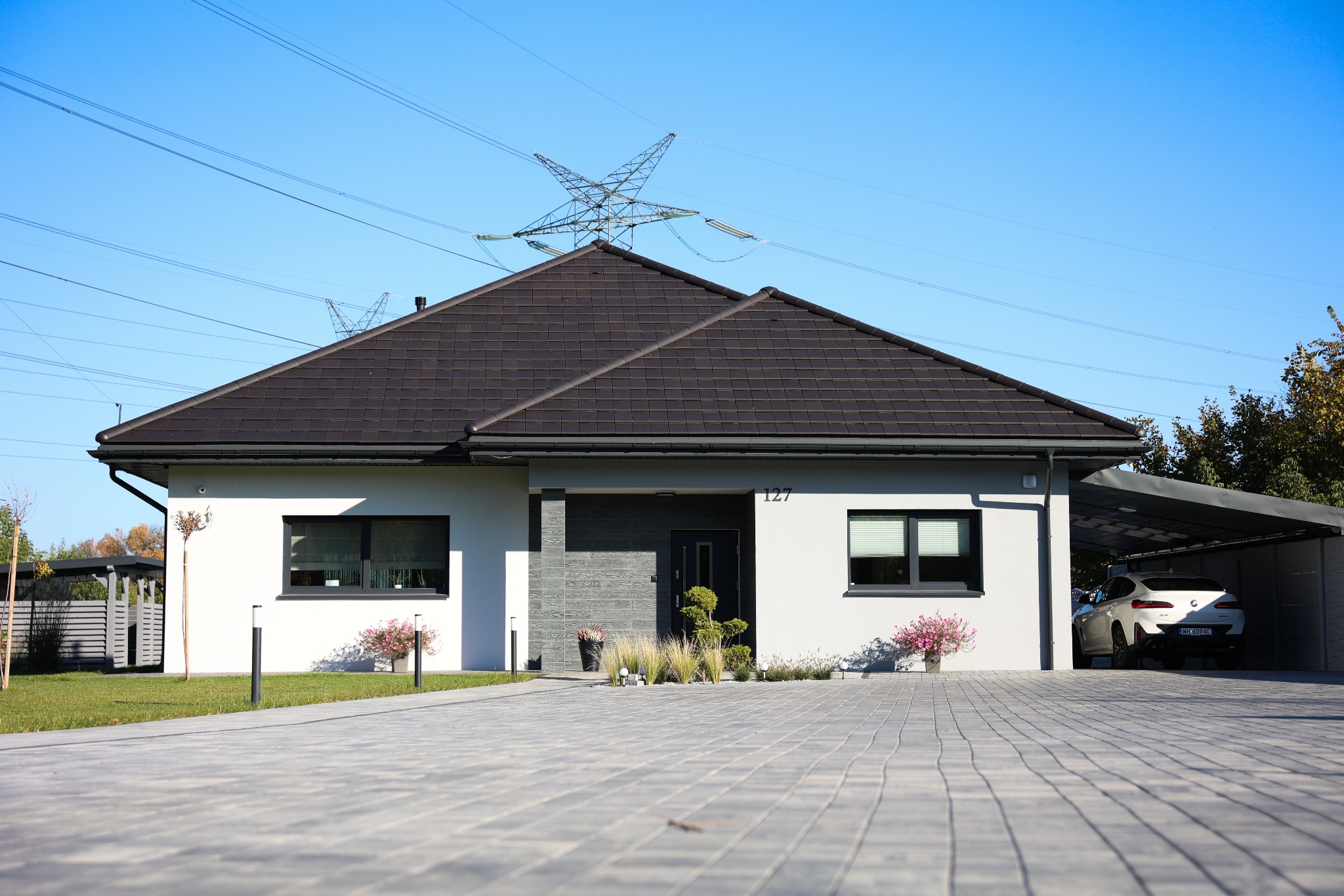 Parterowy dom z ciemnym dachem i jasną elewacją, numer 127, z podjazdem z kostki brukowej i wiatą garażową z białym samochodem. Widok na słupy wysokiego napięcia.