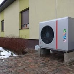 Alkar Professional - Szara pompa ciepła zamontowana na betonowych blokach przy ścianie budynku z brązowymi roletami i resztkami śniegu. Widok z boku, lekko z dołu.