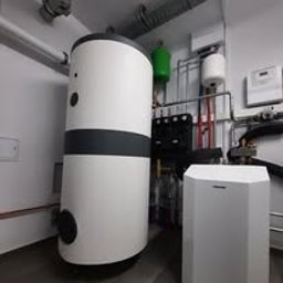 Alkar Professional - Biały zasobnik ciepłej wody i pompa ciepła w kotłowni w Koninie. Widoczne rury instalacji grzewczej i osprzęt. Nowoczesna instalacja w pomieszczeniu.