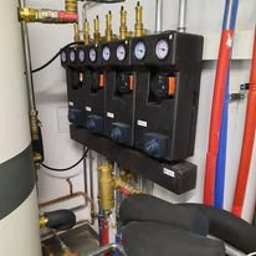 Alkar Professional - Czarny rozdzielacz pompy ciepła zamontowany na ścianie w kotłowni, z widocznymi manometrami, rurami miedzianymi i zbiornikiem. Oświetlenie sufitowe, kąt ujęcia od dołu.